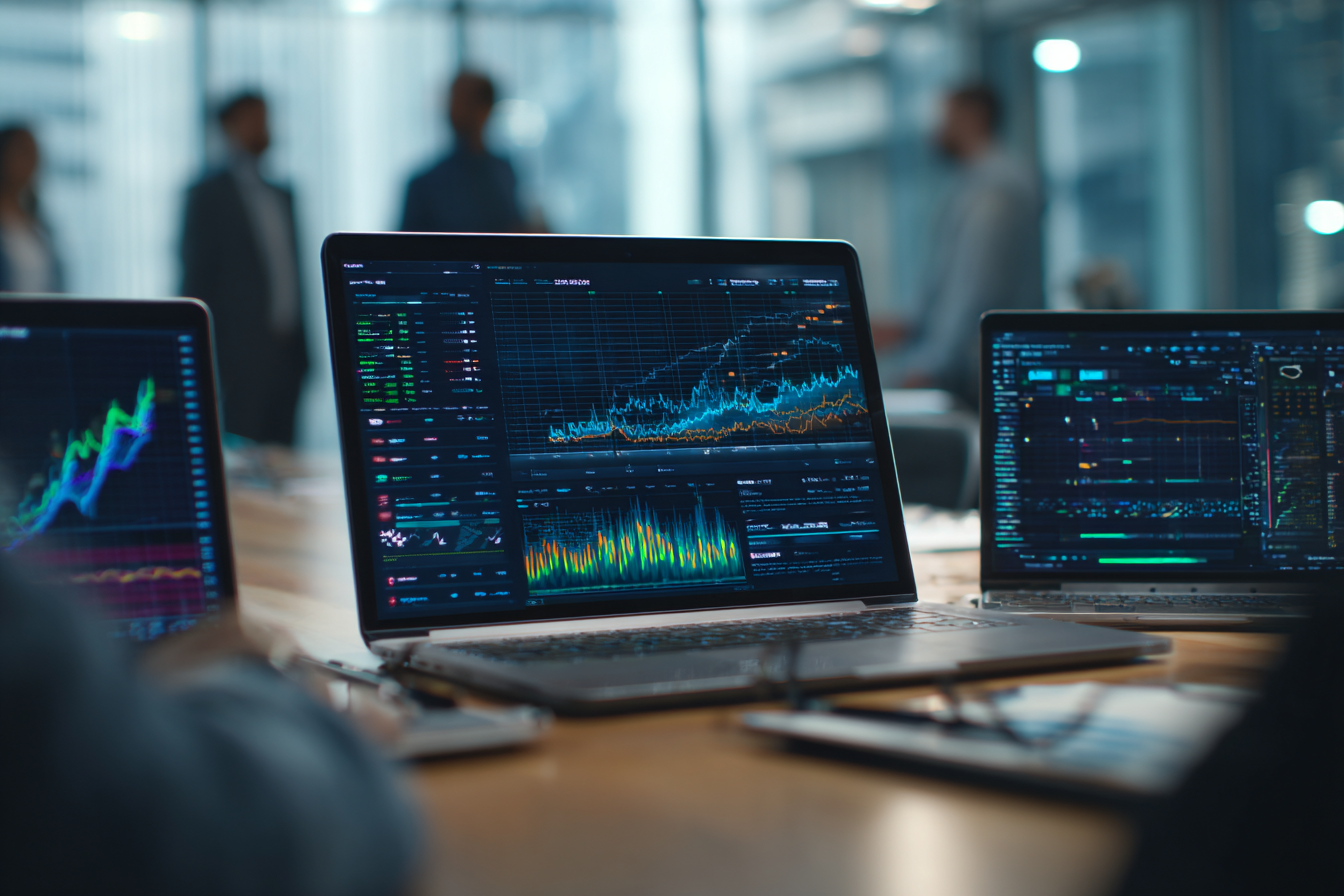 Laptop screens displaying financial graphs and data charts on a meeting table with blurred people in the background.