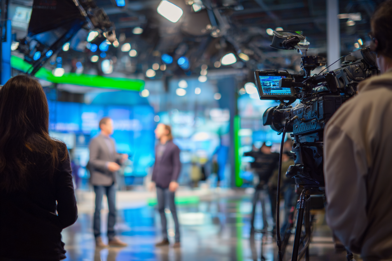 Television studio with camera crew filming two people having a discussion under bright studio lights.
