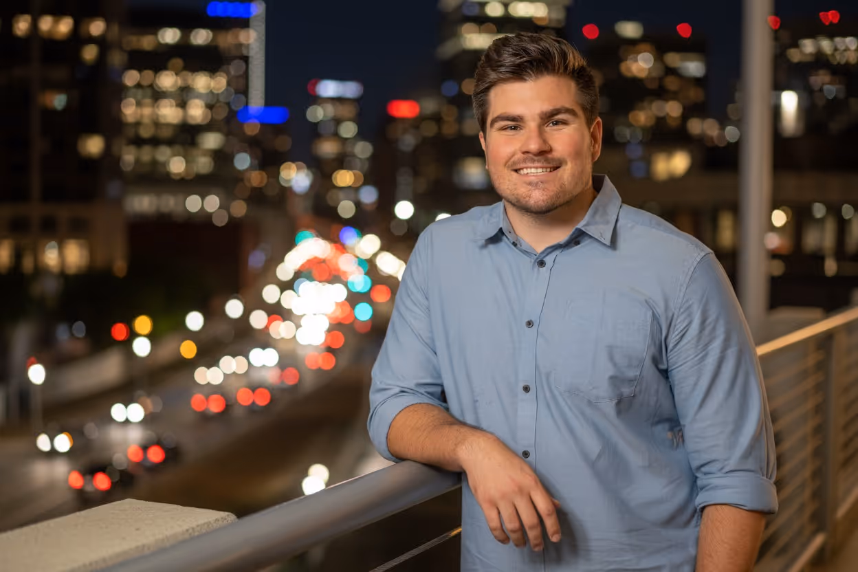 Smiling man in a light blue button-up shirt leaning on a railing with a cityscape and blurred traffic lights at night in the background.
