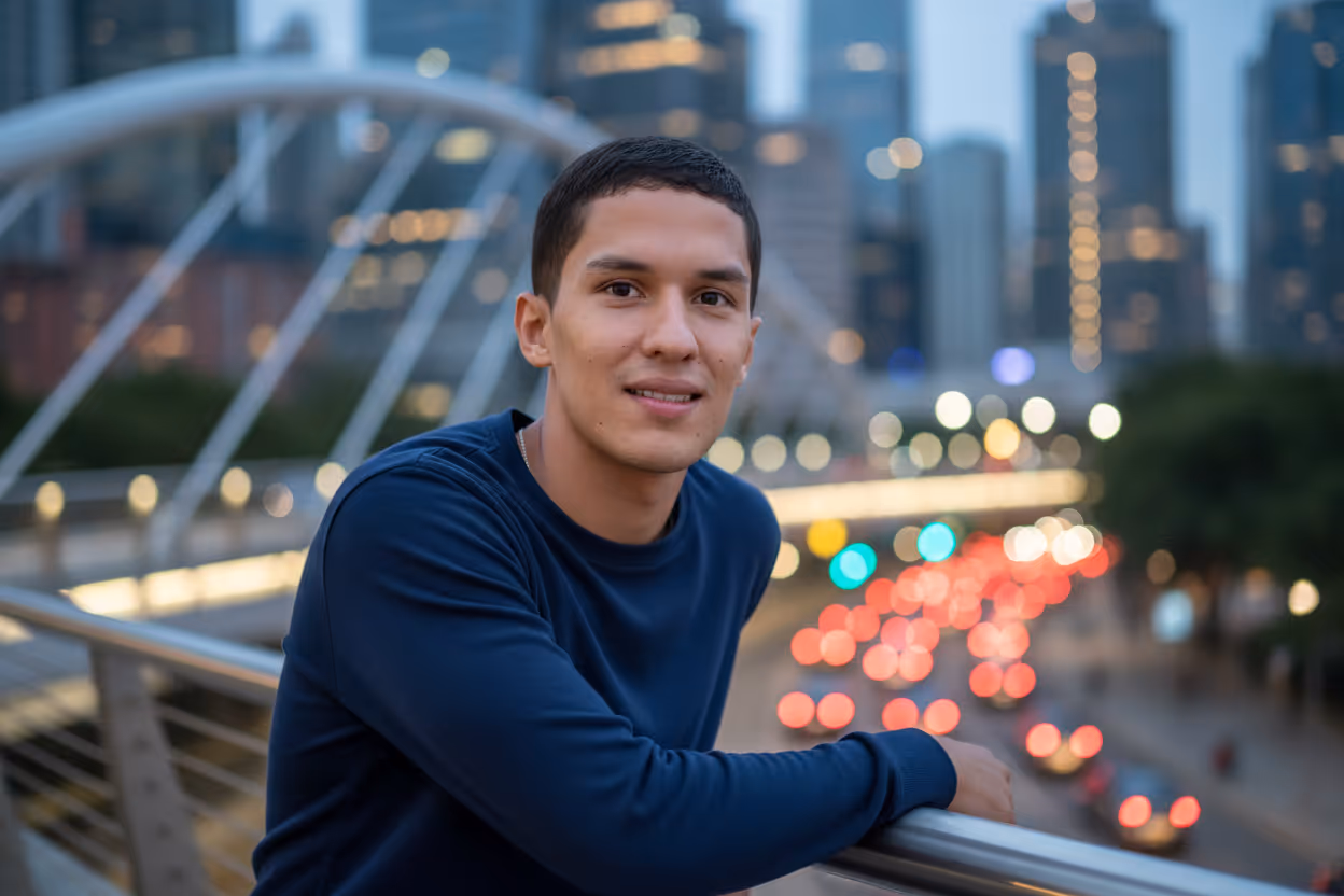 Young man in a navy blue sweater leaning on a railing with a cityscape and blurred traffic lights in the background at dusk.
