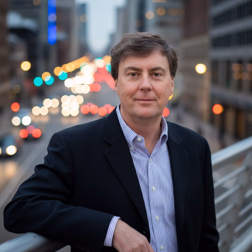 Man in dark blazer and striped shirt leaning on railing with blurred city street and colorful lights in the background.
