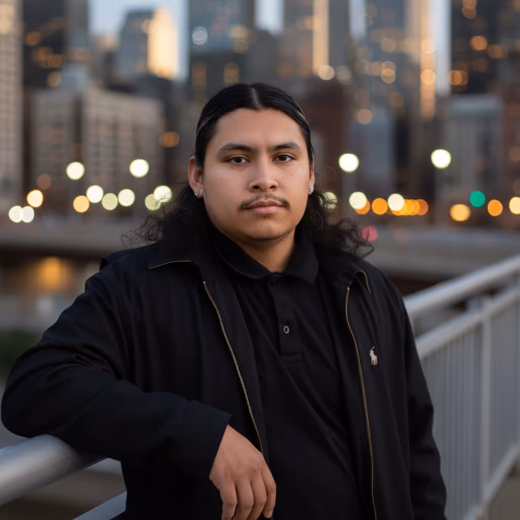 Man with long dark hair and mustache wearing a black jacket and polo shirt, leaning on a railing with a blurred cityscape background at dusk.