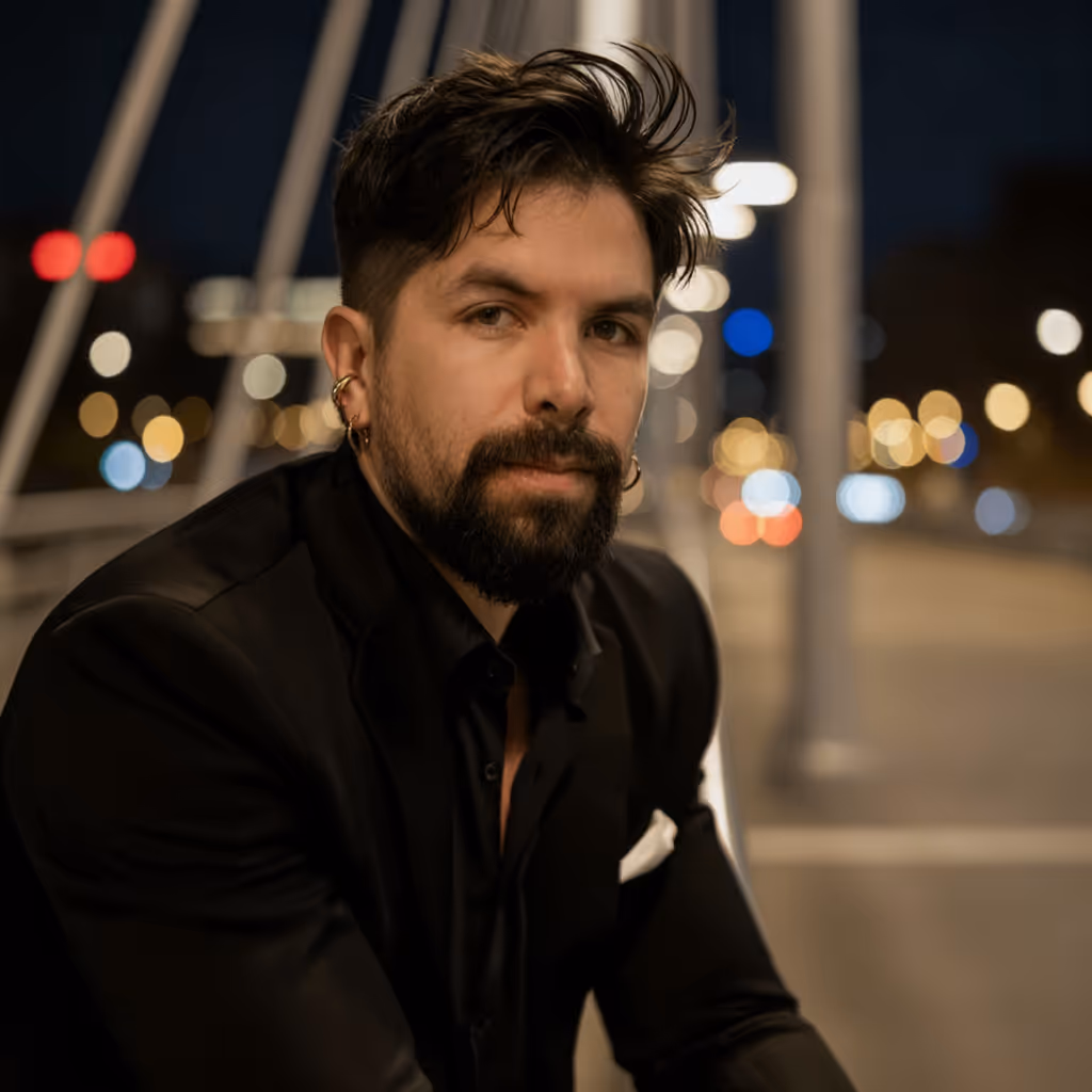Bearded man with dark hair and multiple earrings wearing a black suit jacket, sitting on a bridge at night with blurred city lights in the background.