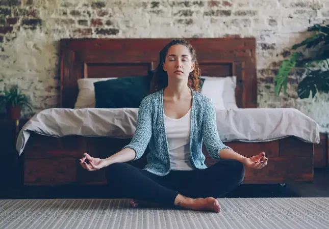 Eine Frau, die im Schneidersitz in einem Schlafzimmer meditiert und den Fokus auf mentale Gesundheit innerhalb der Hormonersatztherapie hervorhebt.