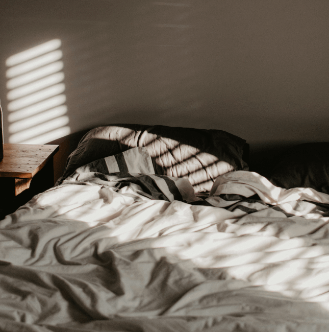 Ein ungemachtes Bett mit durch Jalousien fallenden Sonnenlicht-Schatten, das die Herausforderung eines gestörten Schlafmanagements veranschaulicht.