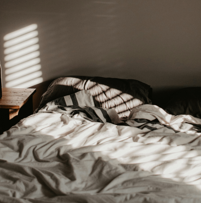 Ein ungemachtes Bett mit durch Jalousien fallenden Sonnenlicht-Schatten, das die Herausforderung eines gestörten Schlafmanagements veranschaulicht.