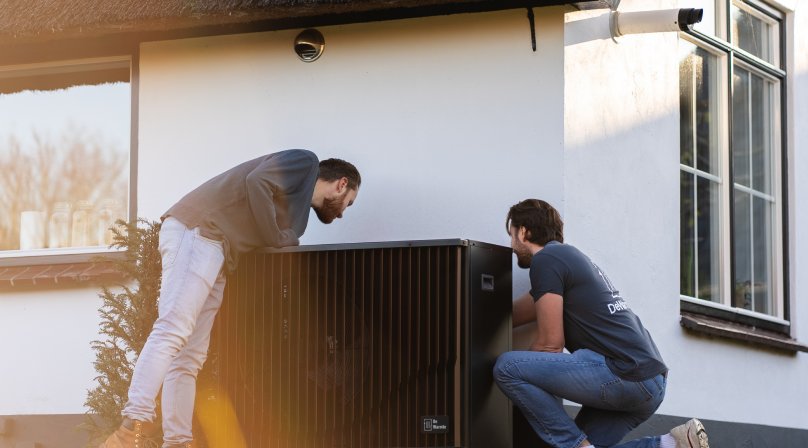 Twee monteurs plaatsen een DeWarmte buitenunit naast de gevel van een woning