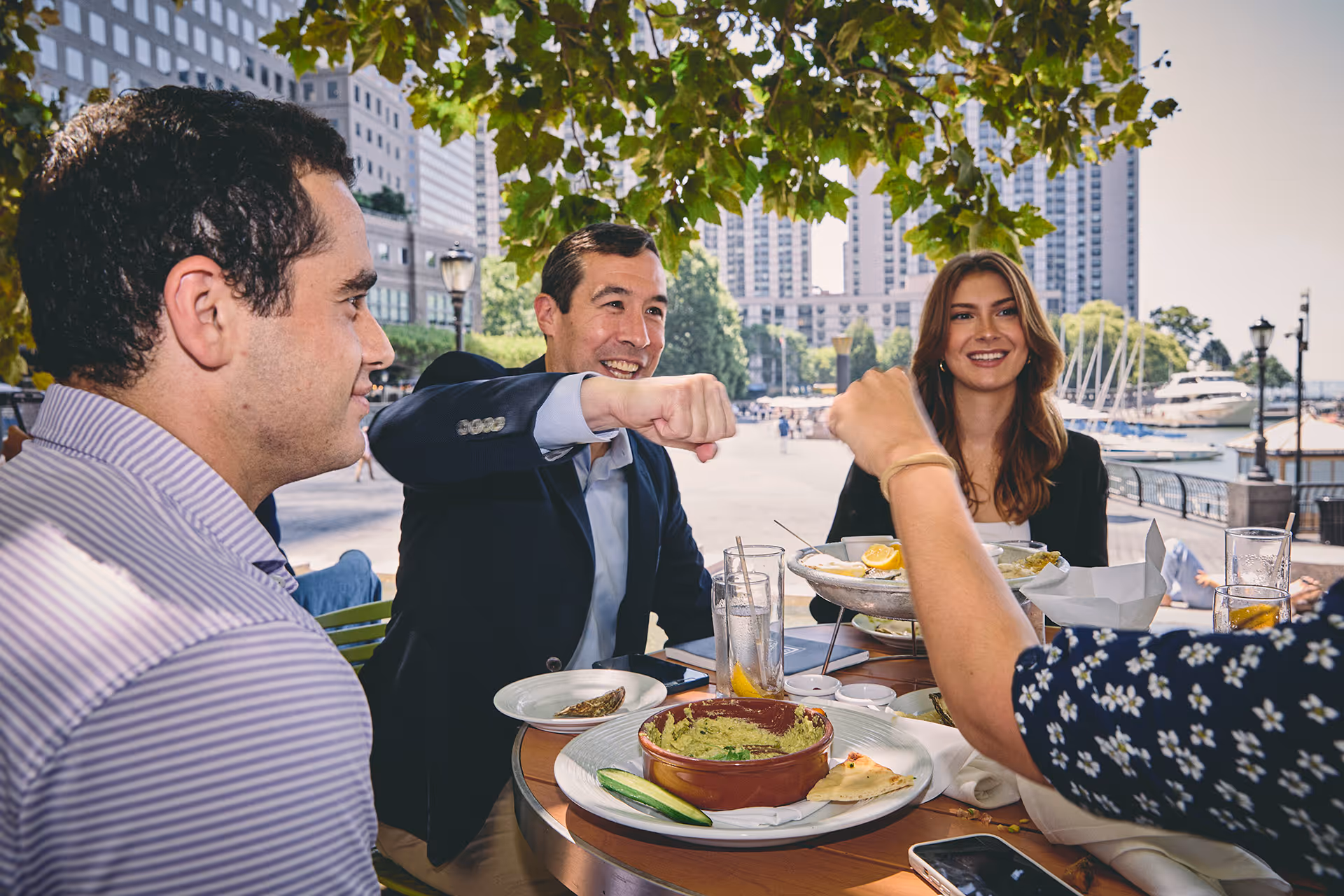 Three people, two men and one woman, sitting at a table outdoors eating food in front of a cityscape with tall buildings and boats in the background.