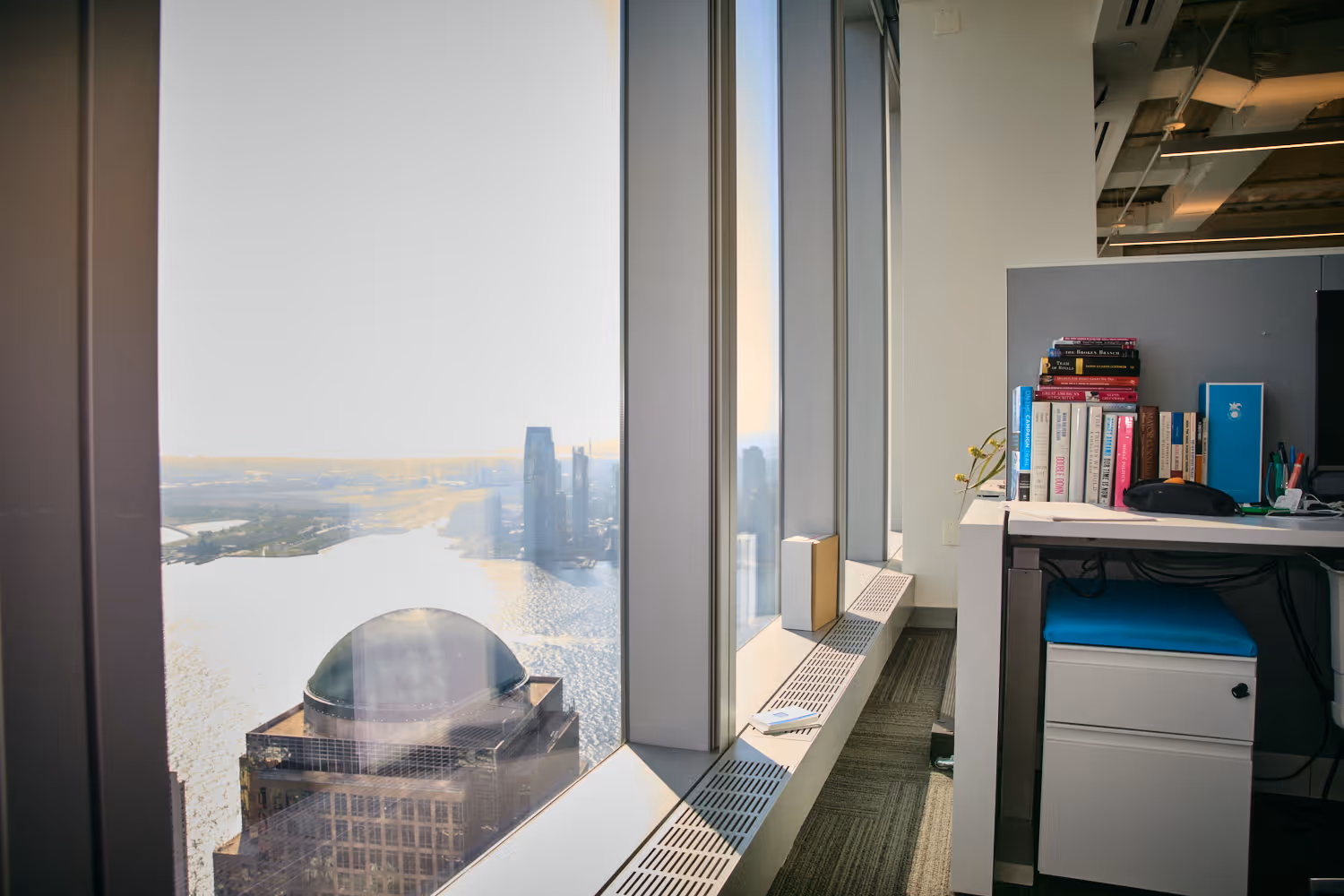 A modern office space with a panoramic view of a city skyline through large windows. The office contains bookshelves, desk, and other office supplies.
