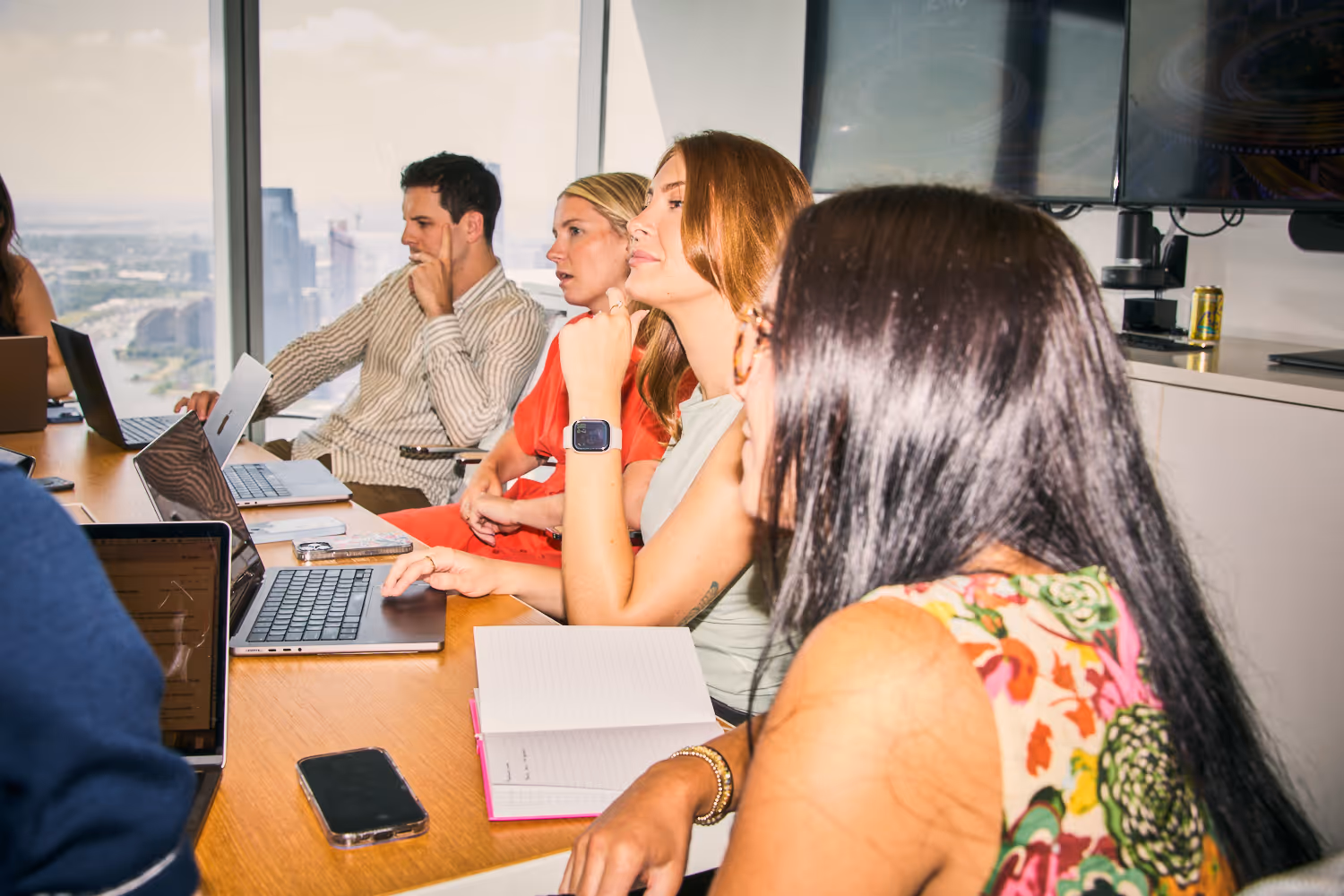 A group of young professionals working together in an office setting with a cityscape visible through the windows behind them.