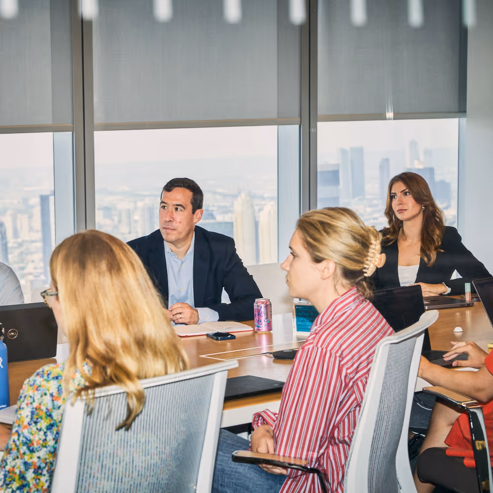 A group of business professionals in a conference room setting, discussing and collaborating on a project. The image shows a meeting room with large windows overlooking a city skyline.
