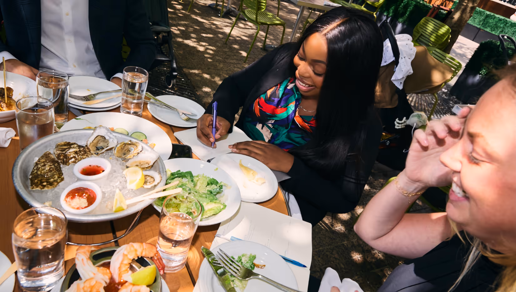 Two women smiling and laughing while seated at an outdoor table with assorted seafood dishes and glasses of water.