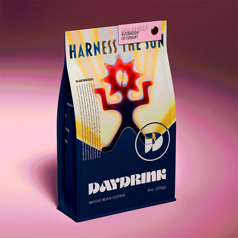 Daydrink 8oz coffee bag in a studio environment with a single light on the bag. The bag is navy blue with a colorful illustration of a gradient Daydrink Sunreacher. The Sunreacher is a character-like logo featuring a sun head with a coffee bean at the center. The color starts from navy blue with a light blue outline around it and ends at the top in red and orange hues. Its arms and legs are in a rigid pose, almost like a statue. The background of the illustration is yellow and orange beams that protrude from behind the Sunreacher, evoking the feeling of the warm sun. Above the Sunreacher, in bold lettering, reads "Harness the Sun". At the bottom right foot of the Sunreacher sits a D in a circle taken from the Daydrink wordmark. Below the illustration sits a Daydrink wordmark with "whole bean coffee" "8 oz" underneath. There is a tag with the name "Colombia-Watermelon Co-ferment" attached to the top right corner of the bag with a metal screw.