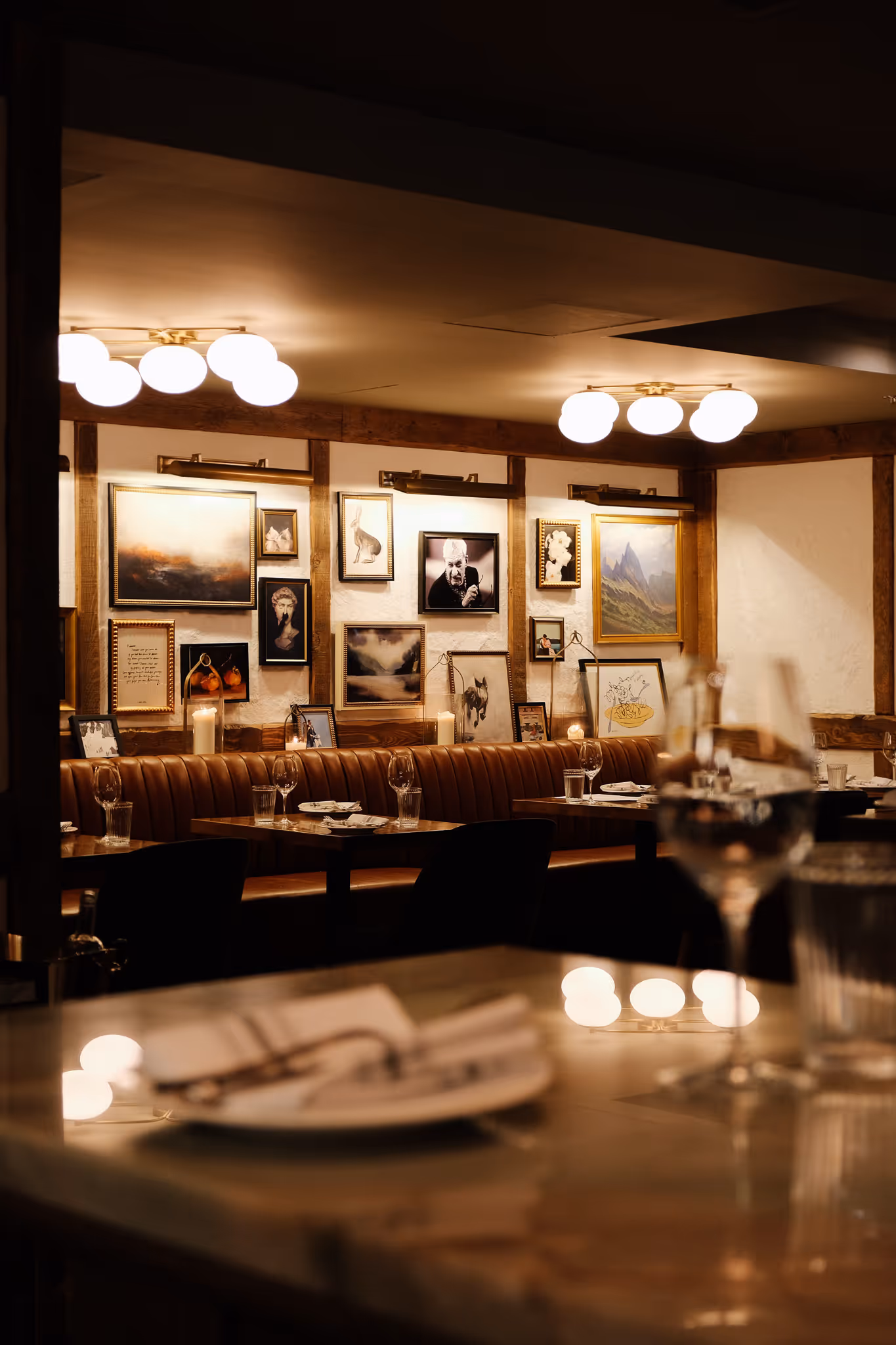 Cozy restaurant interior with brown leather seating, wooden tables set with glasses and cutlery, and a wall decorated with framed paintings and photos under soft ceiling lights.