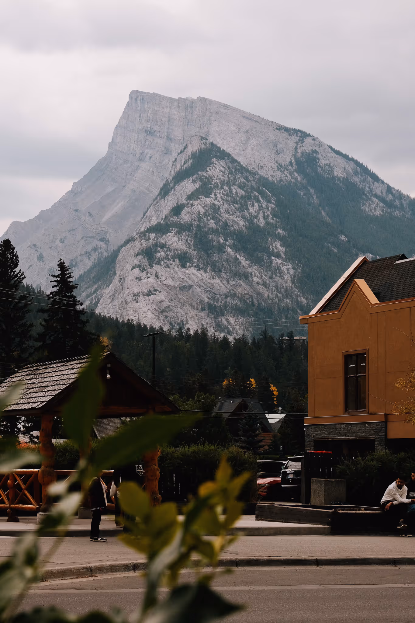 Large rocky mountain with pine trees at its base behind a small town area with wooden structures and a yellow building.