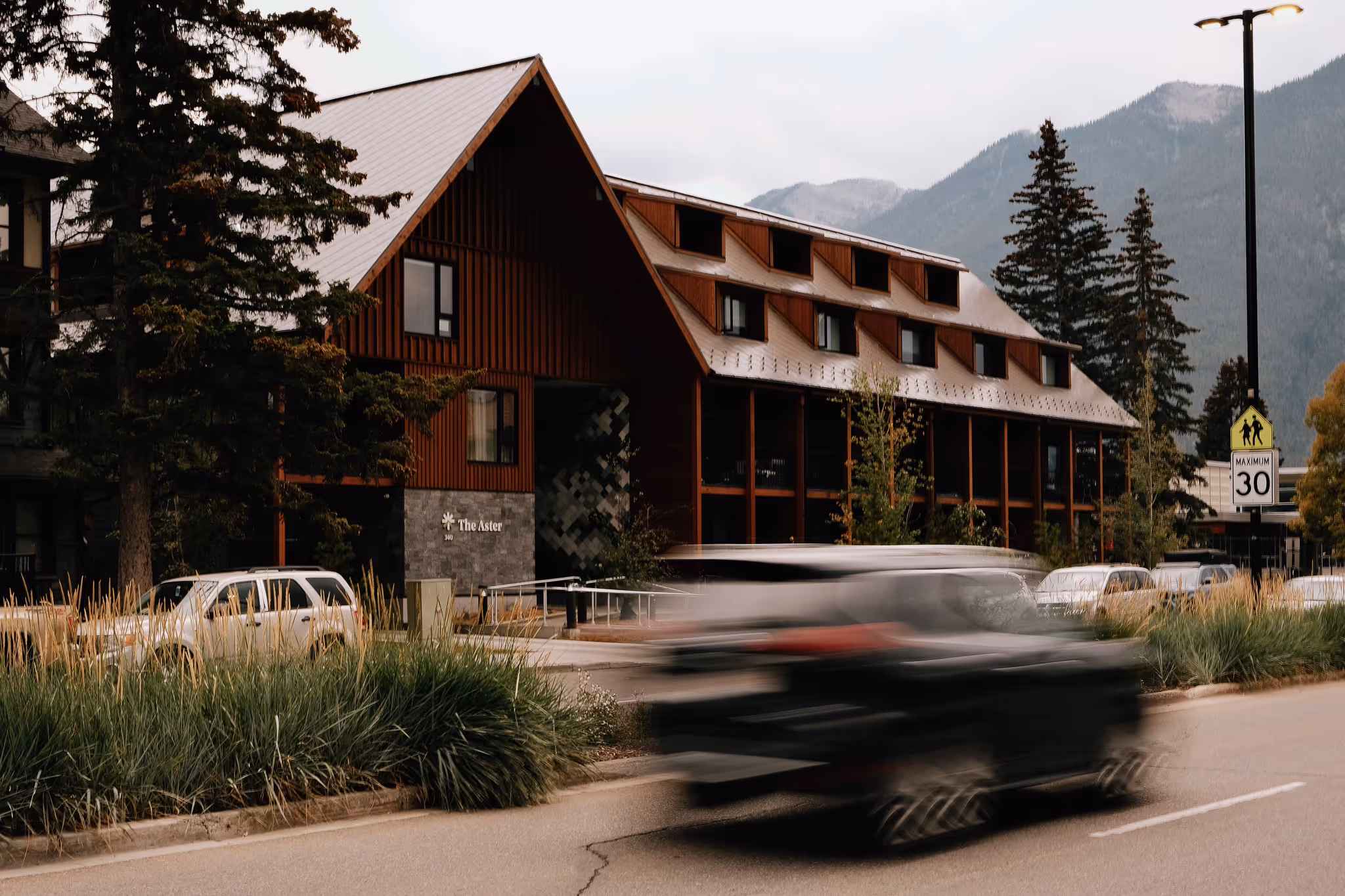 The Aster building with a steep triangular roof beside a road with blurred black vehicle and parked cars, with mountains and trees in the background.