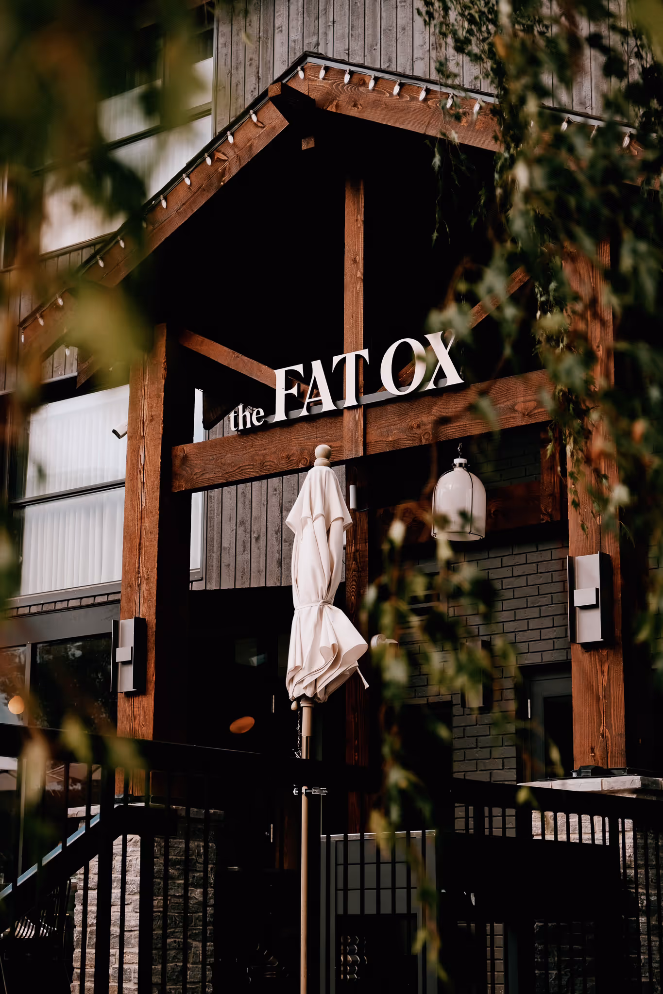 Wooden entrance structure of The Fat Ox restaurant with a folded white umbrella in front and blurred foliage partially obscuring the view.