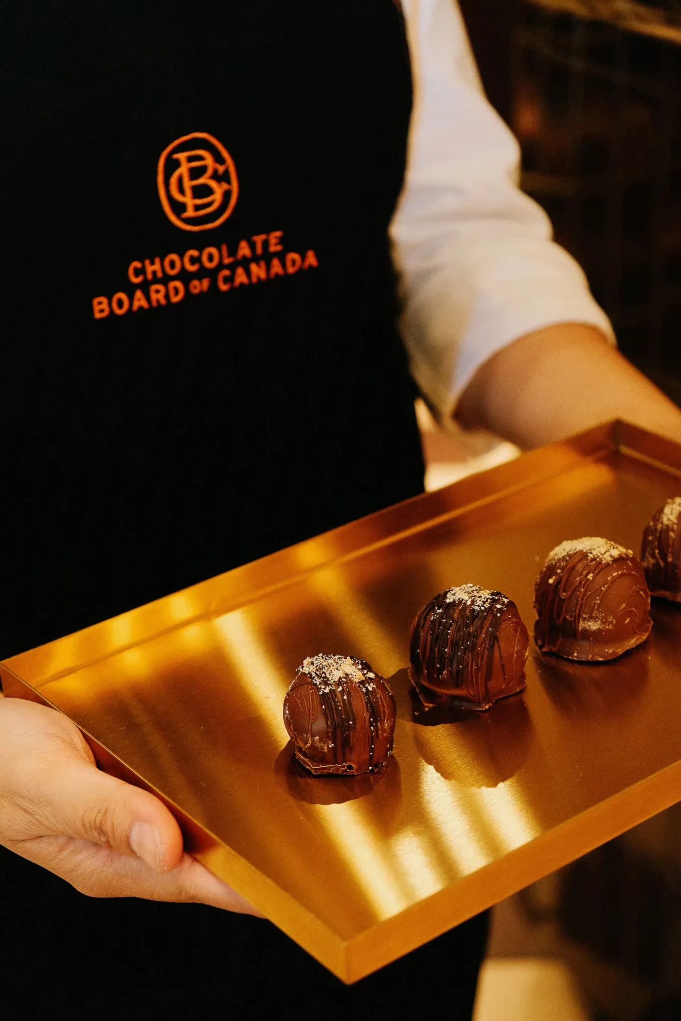 Person wearing a Chocolate Board of Canada apron holding a gold tray with four decorated chocolate truffles.