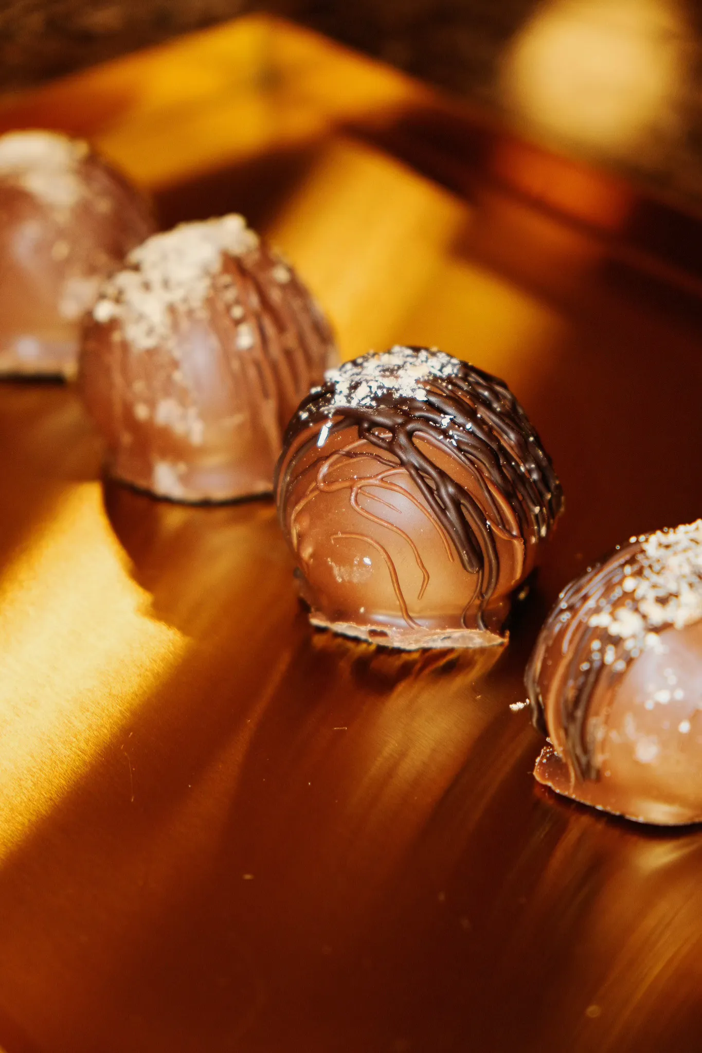 Close-up of four chocolate truffles with decorative drizzles and sprinkled toppings on a reflective golden surface.