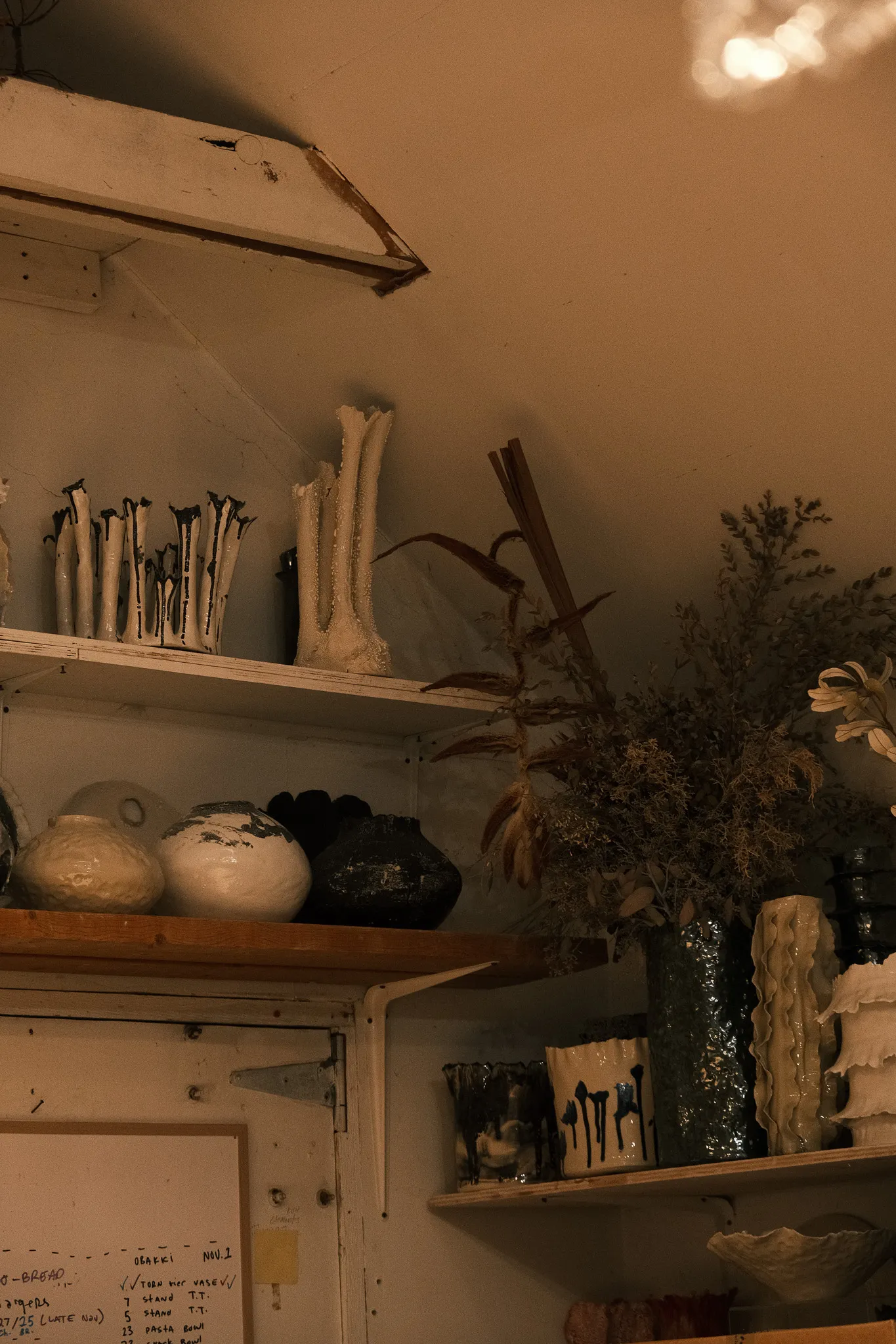 Shelves with a collection of various ceramic vases and pottery, along with dried plants in a tall green vase in a dimly lit room.