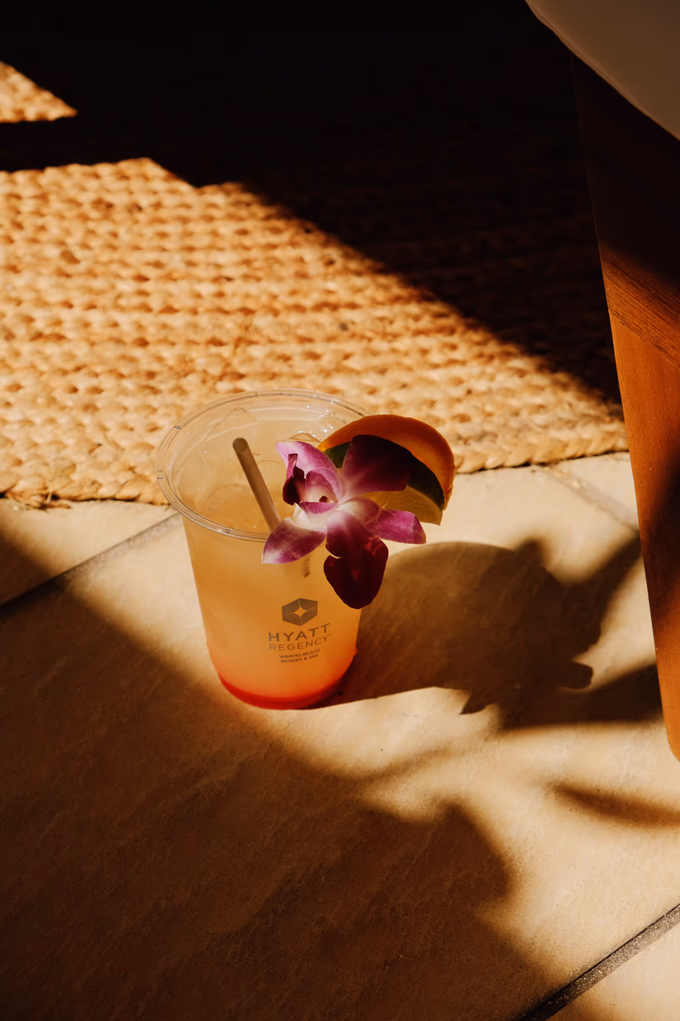 Cold tropical drink in a clear plastic cup with a straw, decorated with a purple orchid and citrus slices, casting a shadow on the floor.