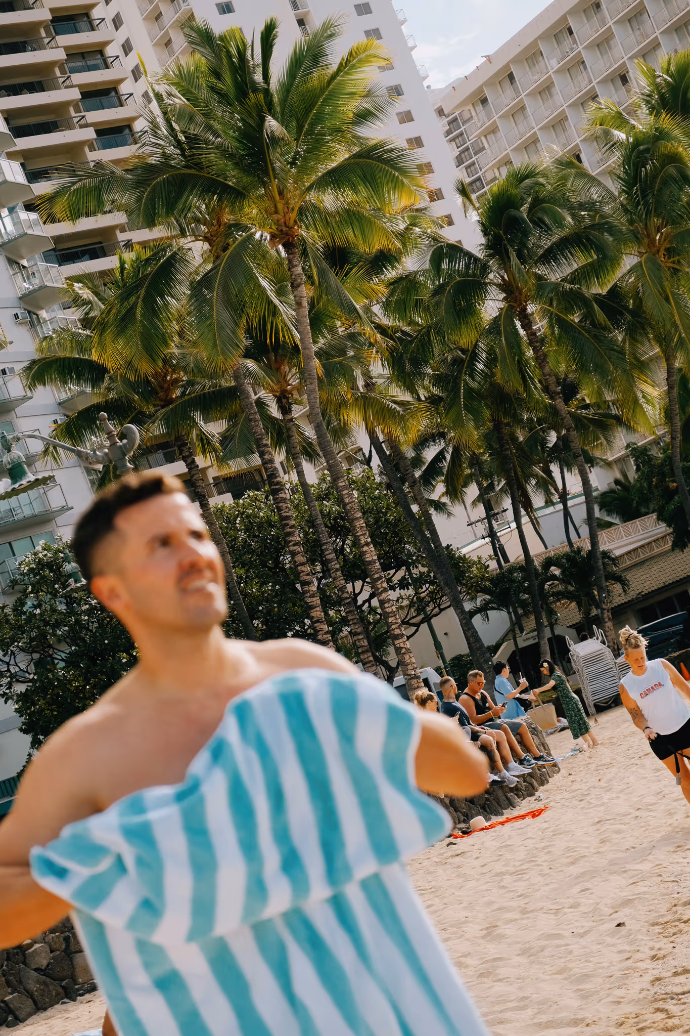 Blurred man holding a blue and white striped towel on a sandy beach with palm trees and tall buildings in the background.