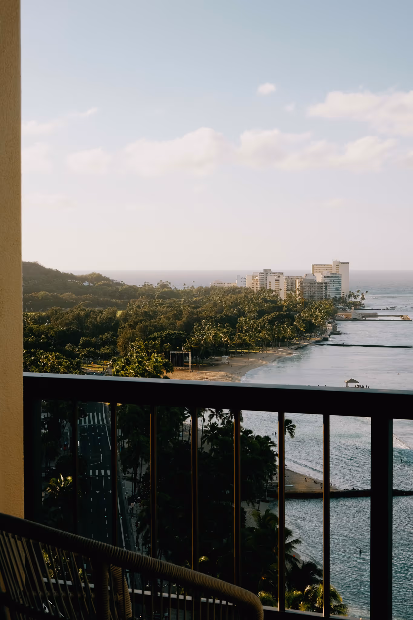 View of a coastal city beach lined with palm trees and buildings, seen from a balcony with railing.