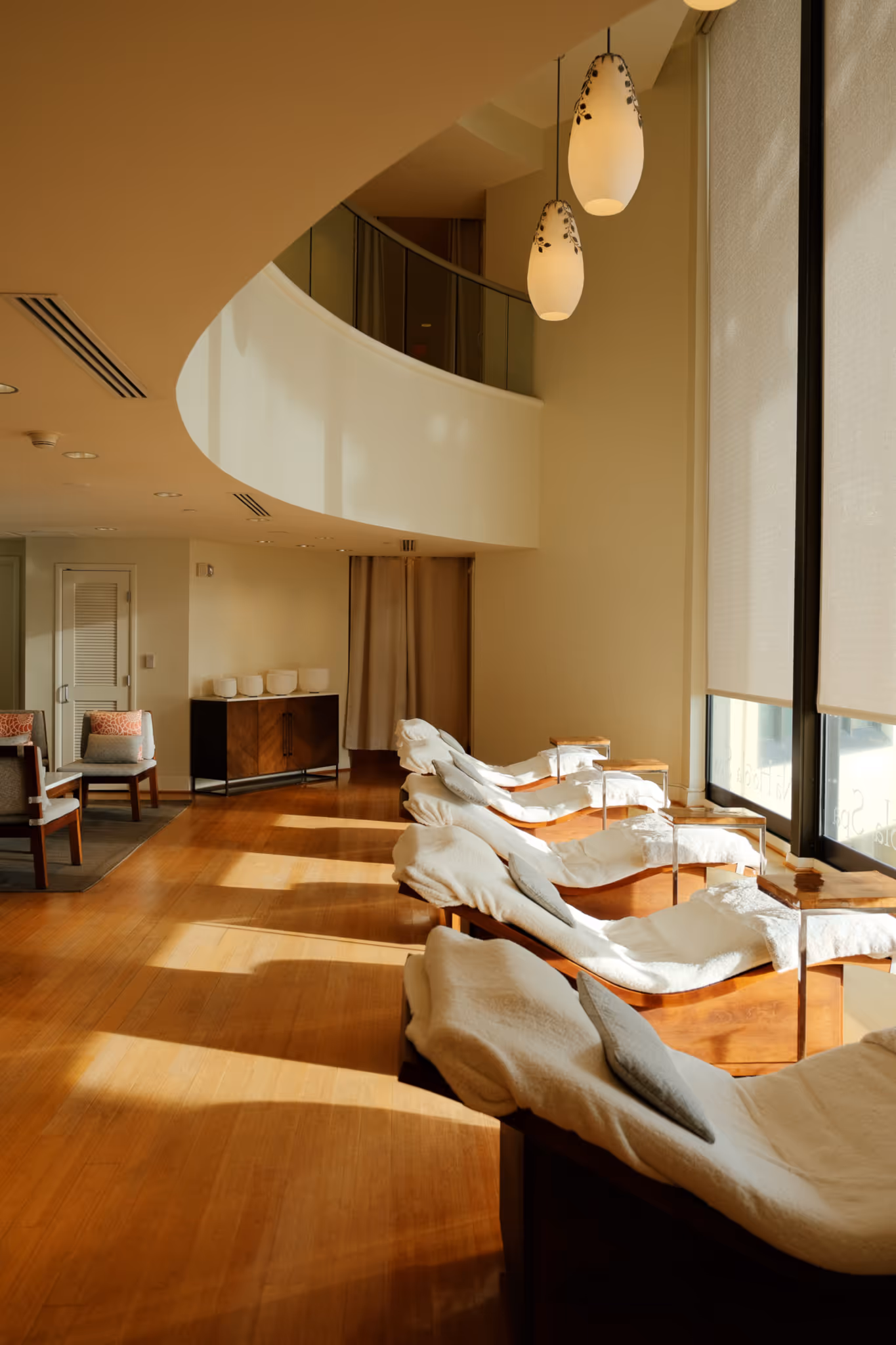 Sunlit modern spa lounge with cushioned chaise lounges lined up next to large windows covered with blinds.