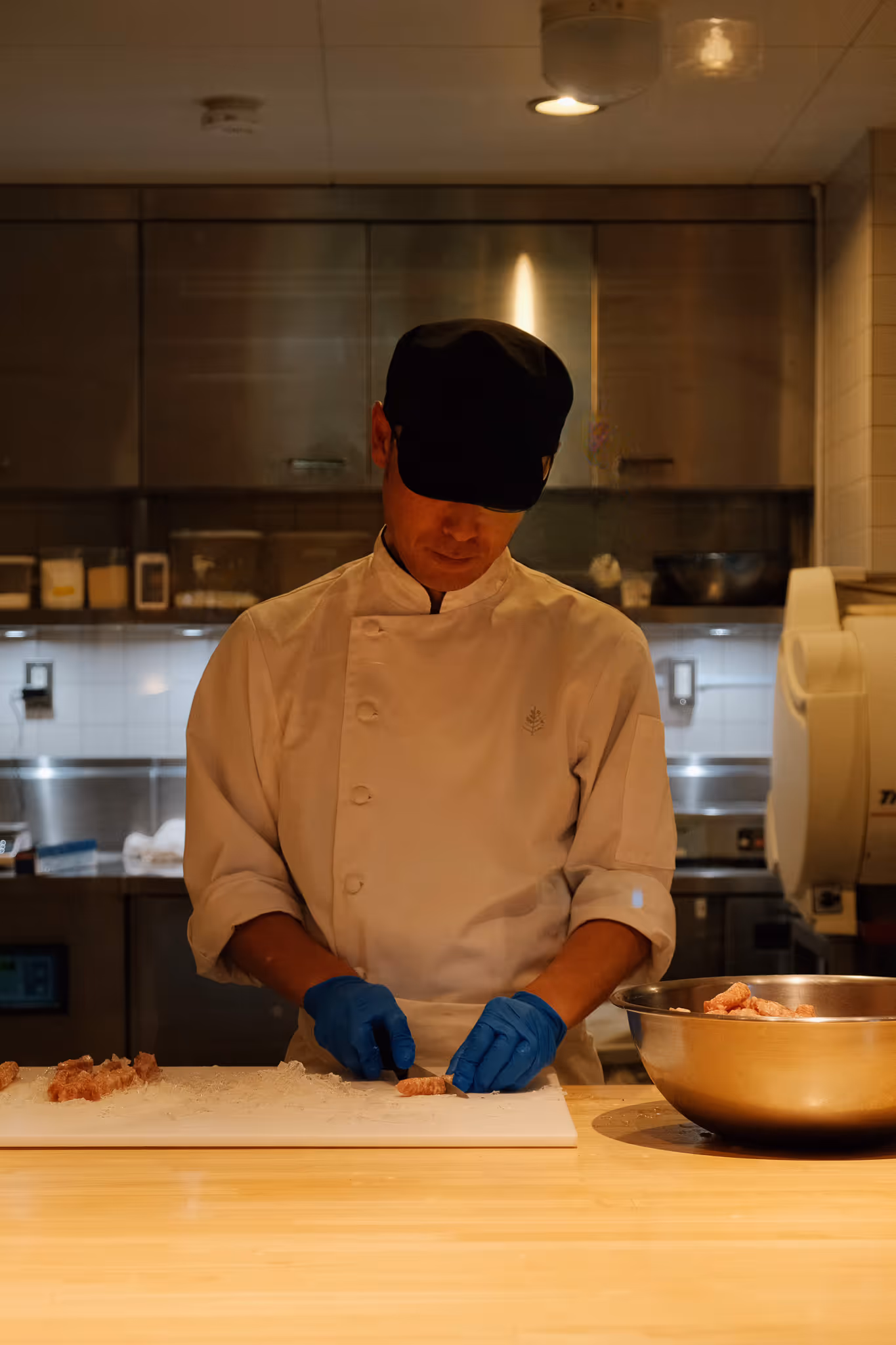 Chef wearing a black cap and white uniform, cutting food on a white board in a professional kitchen.