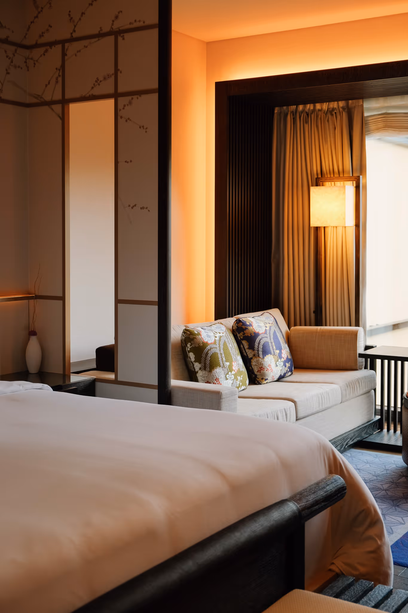 Cozy hotel room with a large bed, beige sofa with floral pillows, warm lighting, and sliding decorative panels.
