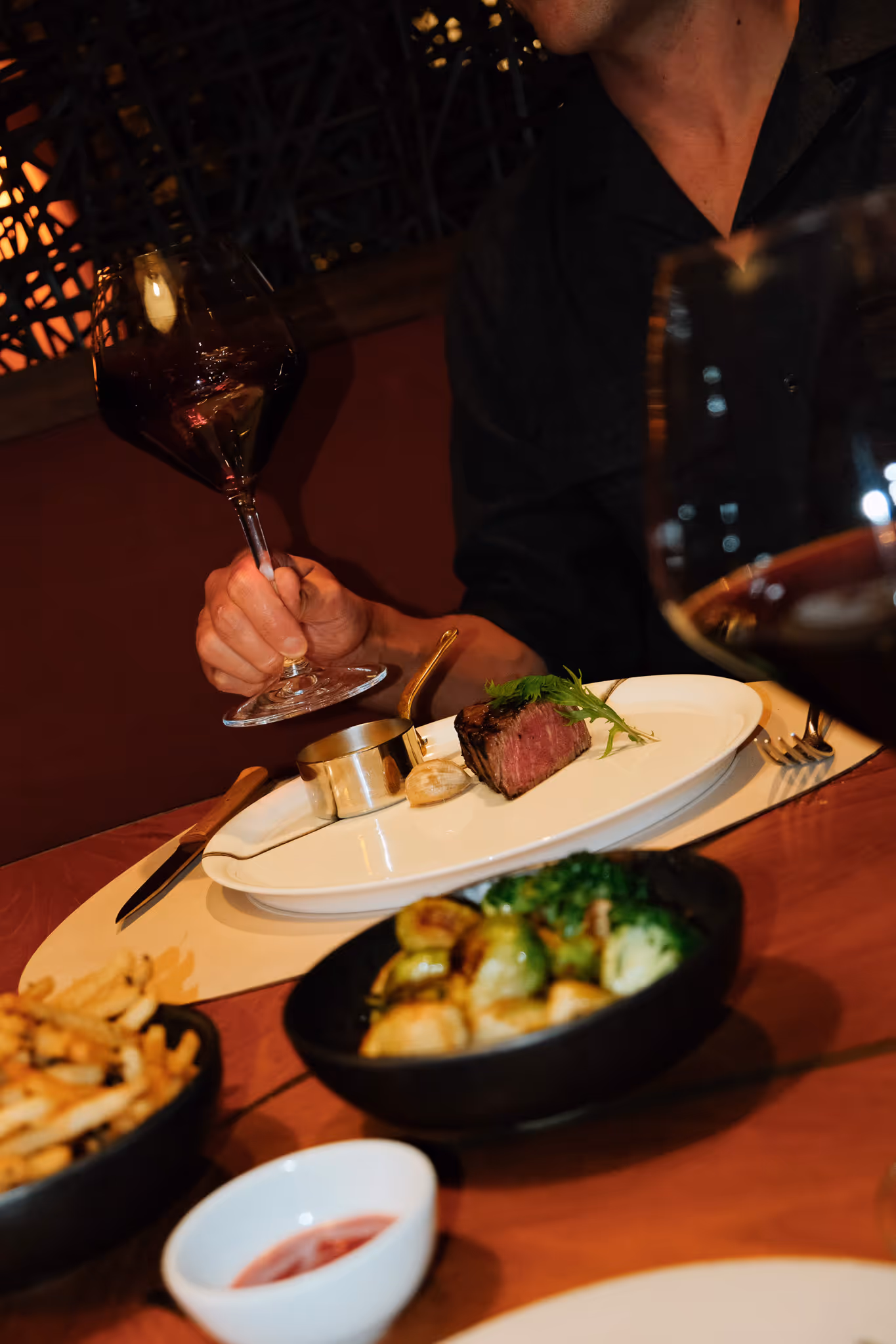 Person holding a glass of red wine at a table with a plated steak, roasted vegetables, fries, and dipping sauce.