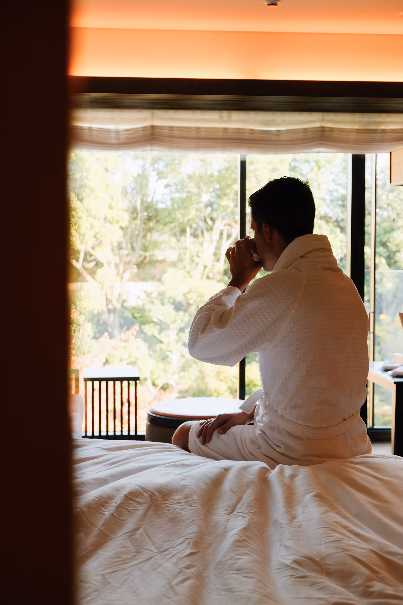 Man in a white bathrobe sitting on a bed, drinking from a cup and looking out a large window at green trees.