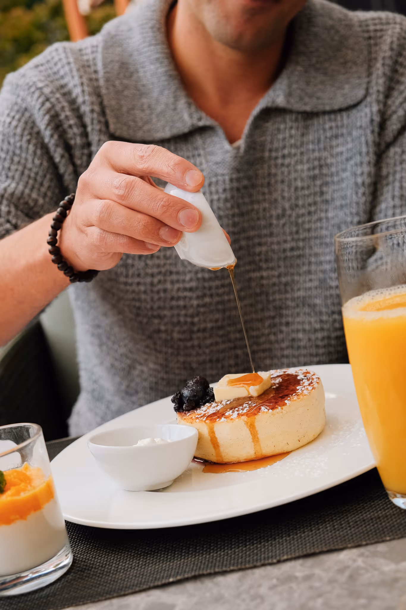 Person pouring syrup over a thick pancake topped with butter and berries on a white plate, with a glass of orange juice nearby.