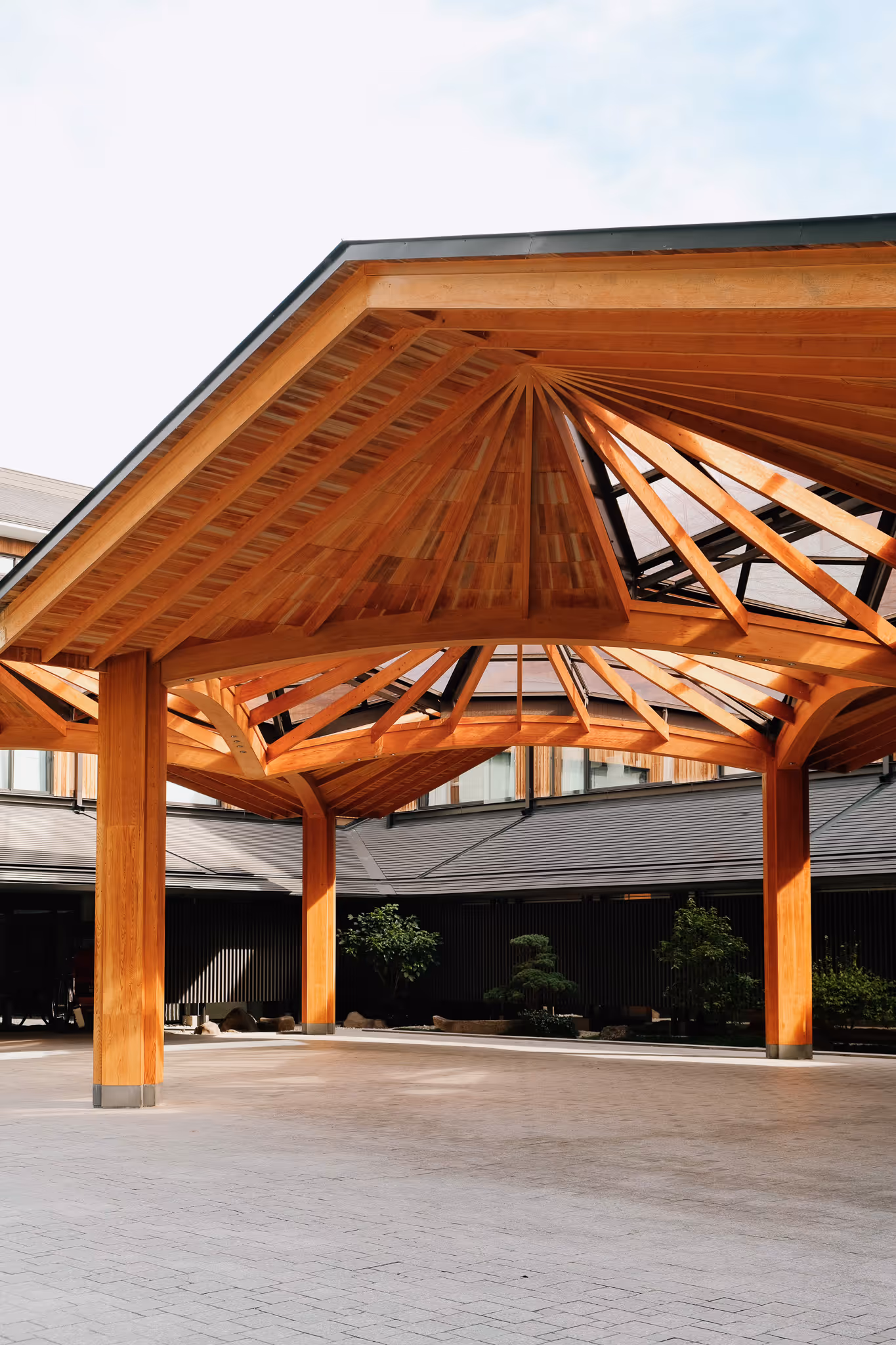 Wooden pavilion with intricate triangular roof beams and a spacious paved floor, surrounded by greenery and modern buildings.