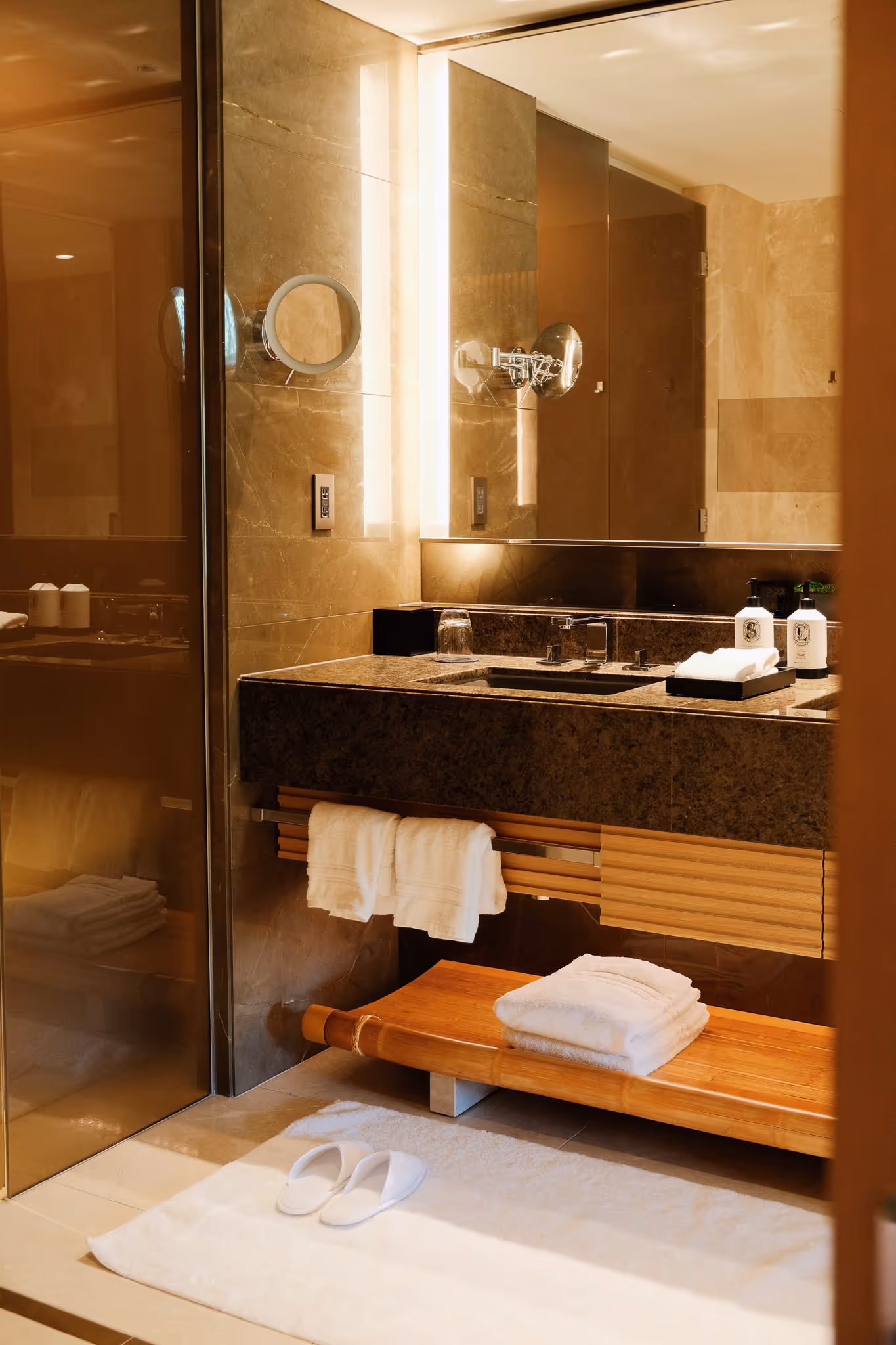 Modern bathroom sink area with granite countertop, towels, slippers on white rug, and illuminated mirror.