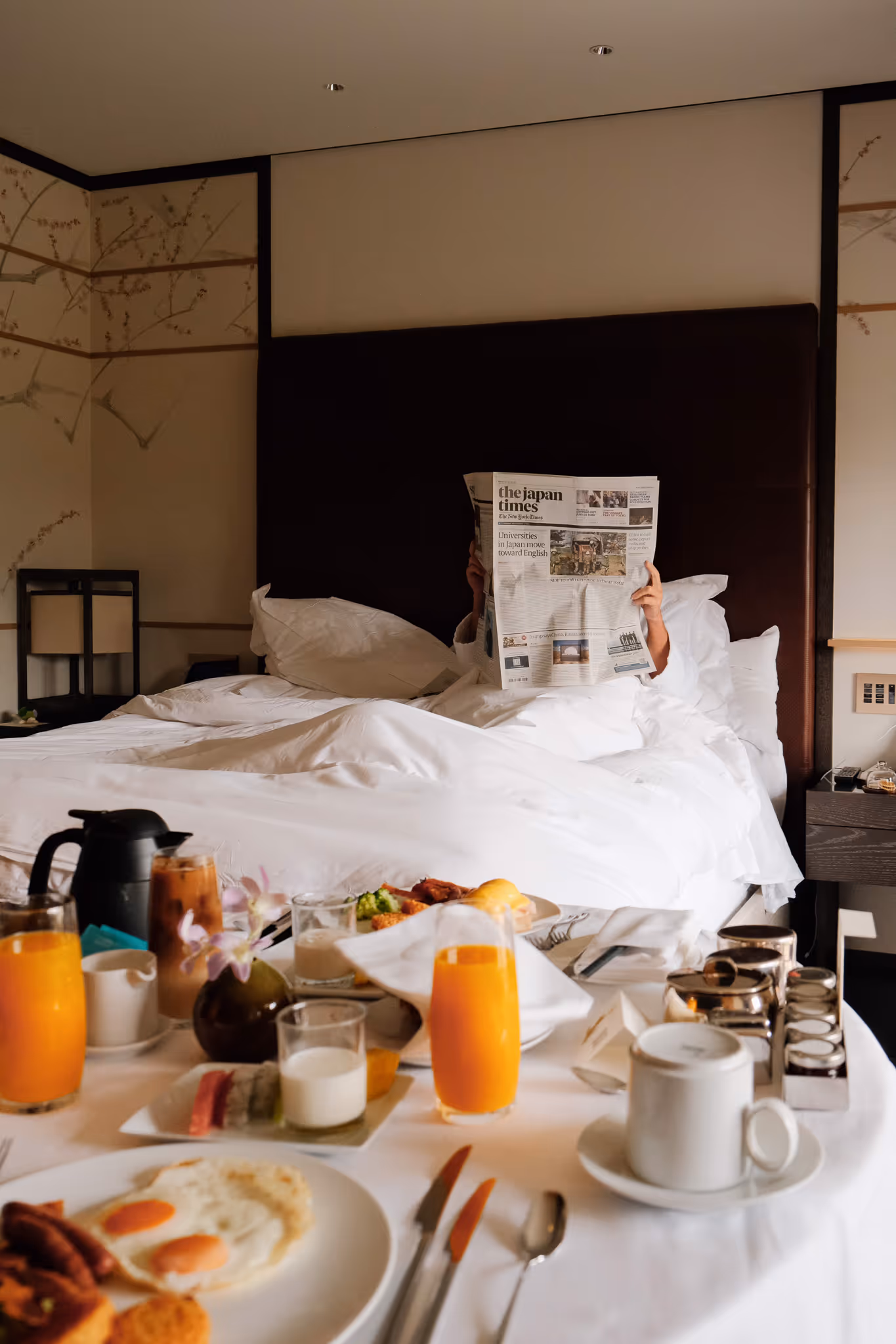 Person in bed reading The Japan Times newspaper with breakfast including eggs, orange juice, and coffee served on a table.