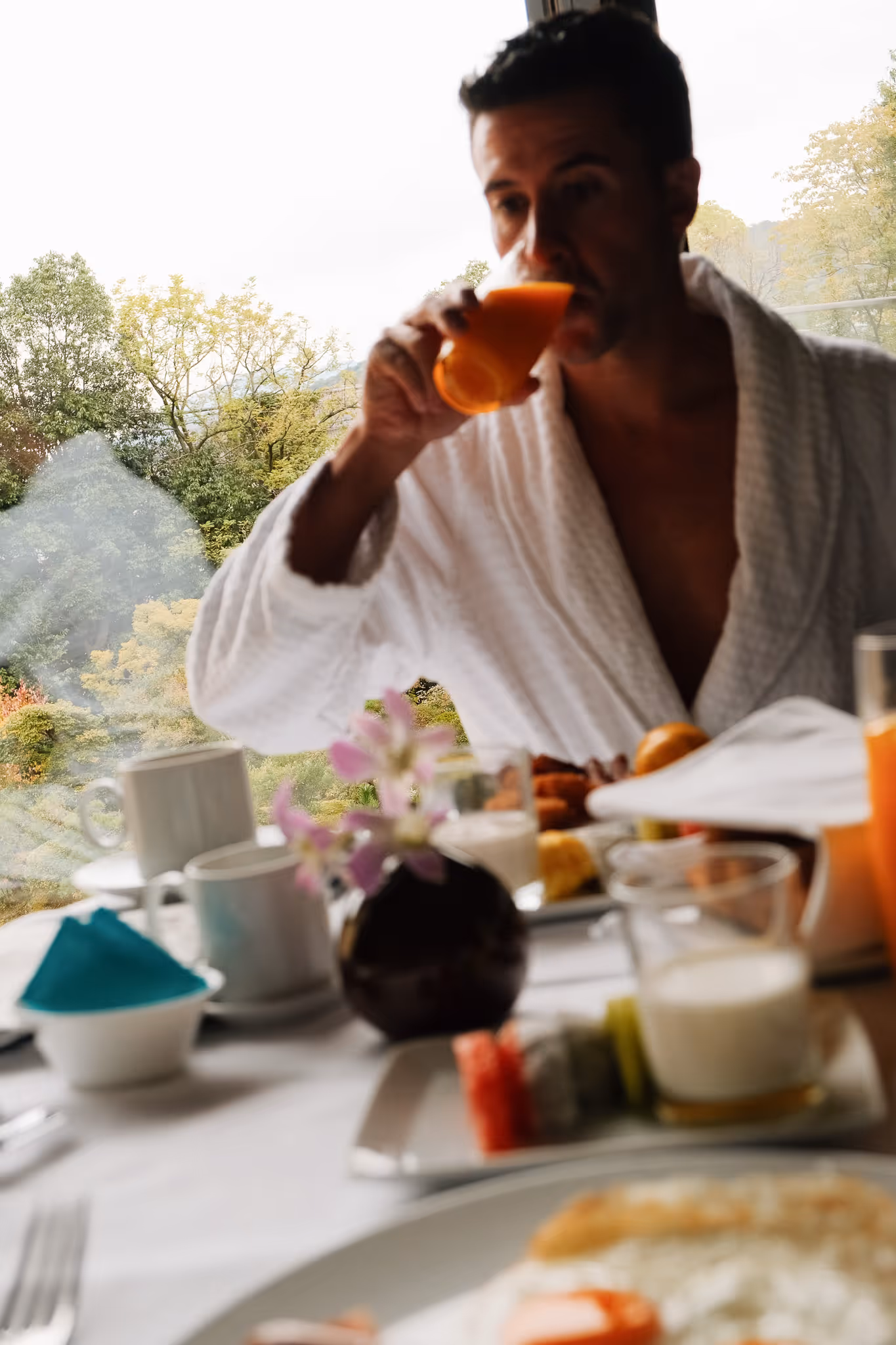 Man in a white bathrobe drinking orange juice at a table set with breakfast food near a window overlooking trees.