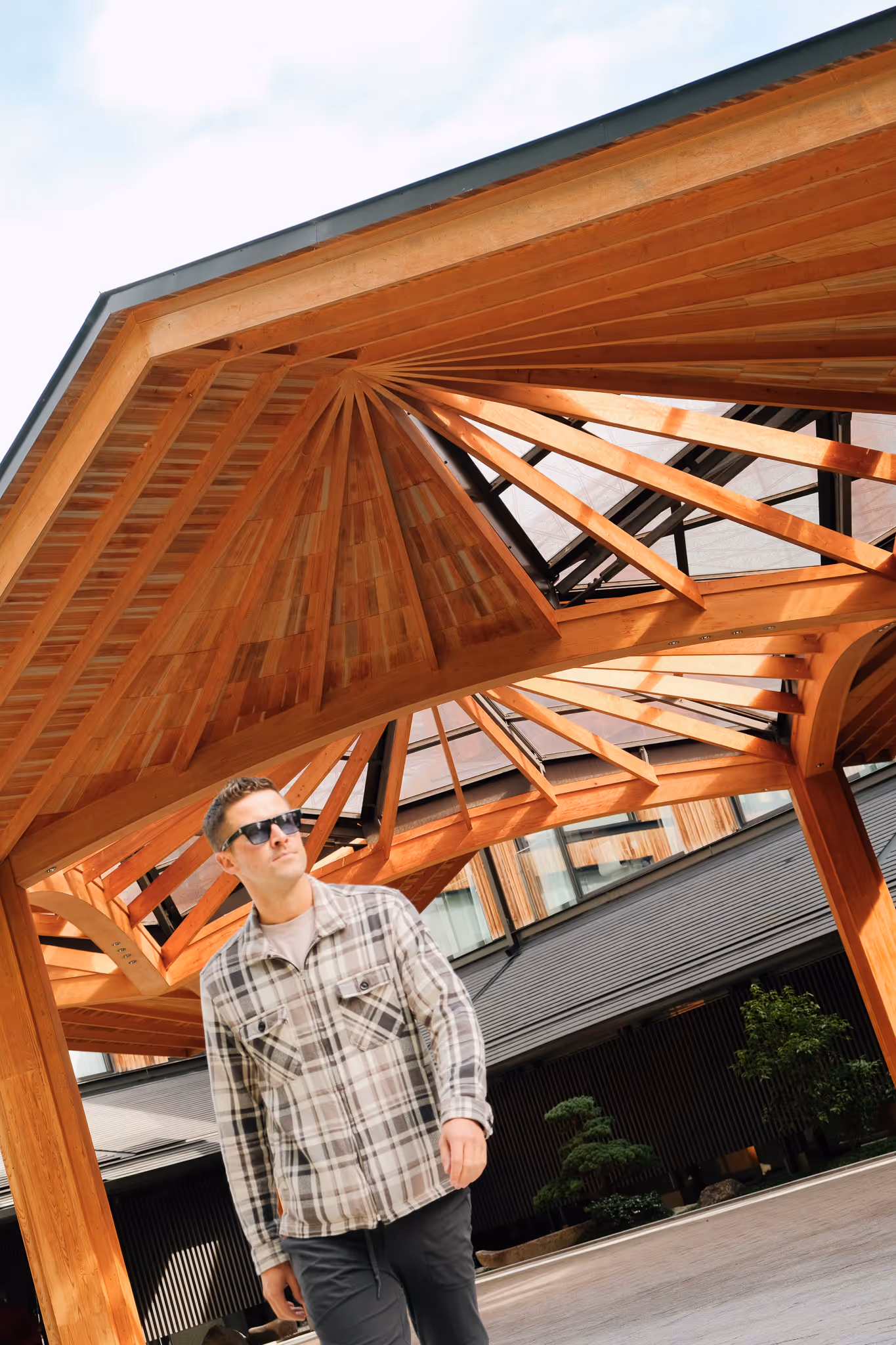 Man in sunglasses walking under a modern wooden pavilion with geometric roof beams and a building in the background.