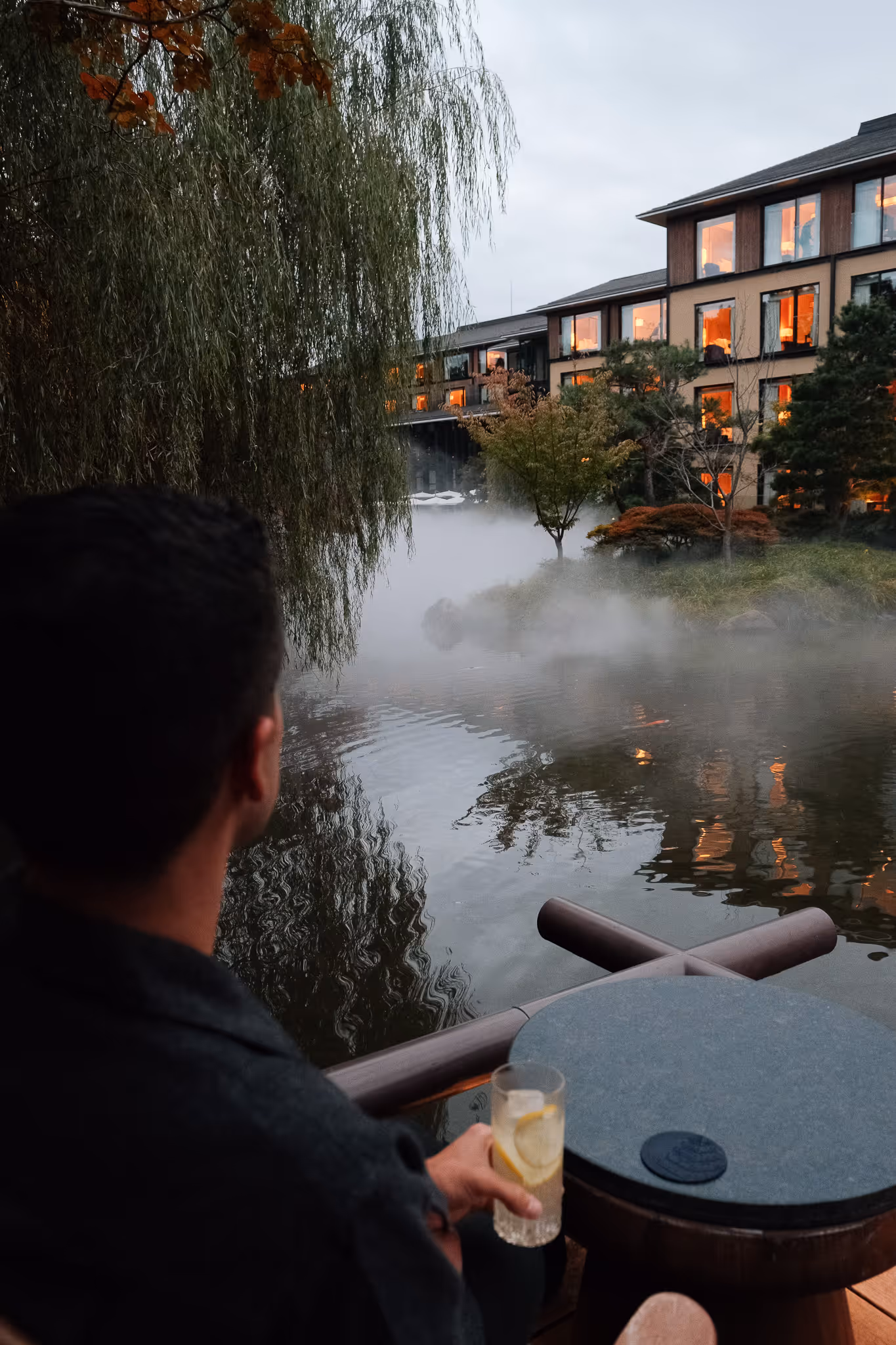 Person holding a glass with lemon slices, sitting by a misty pond with trees and a lit building in the background.