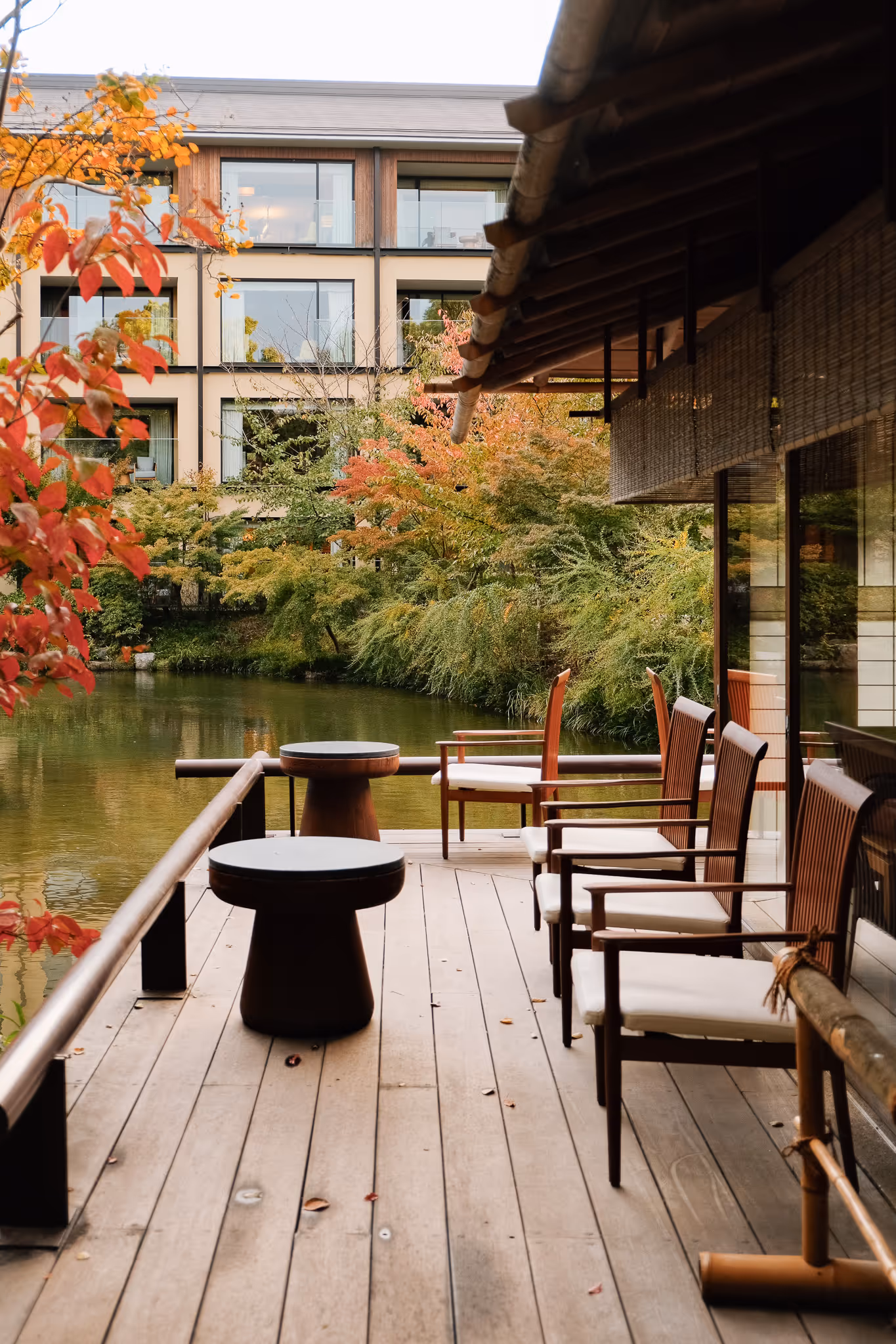 Wooden deck with chairs and small tables overlooking a serene pond surrounded by autumn trees and a modern building.