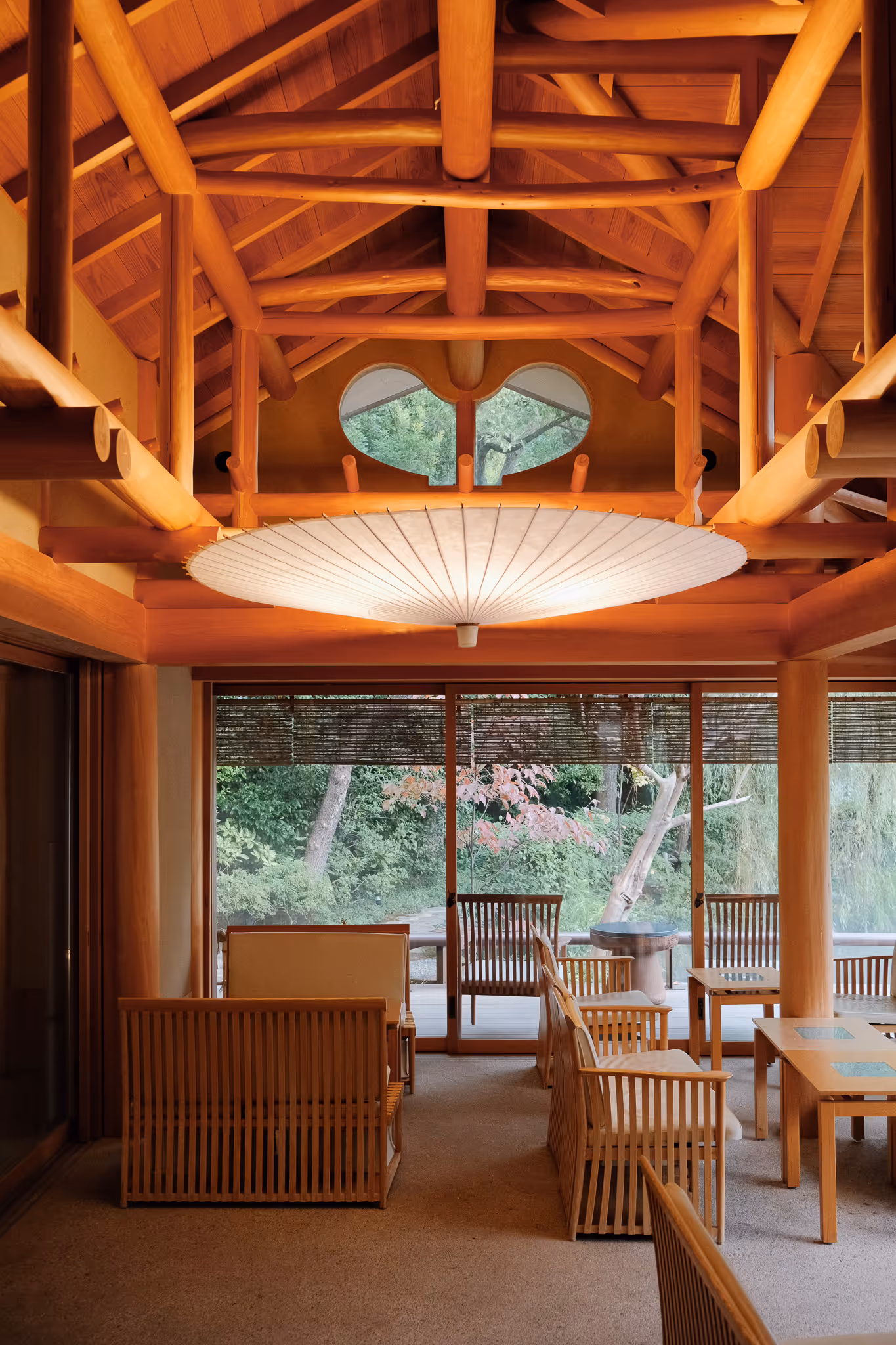 Cozy seating area with wooden chairs and tables inside a room featuring exposed timber beams and large windows overlooking green foliage.