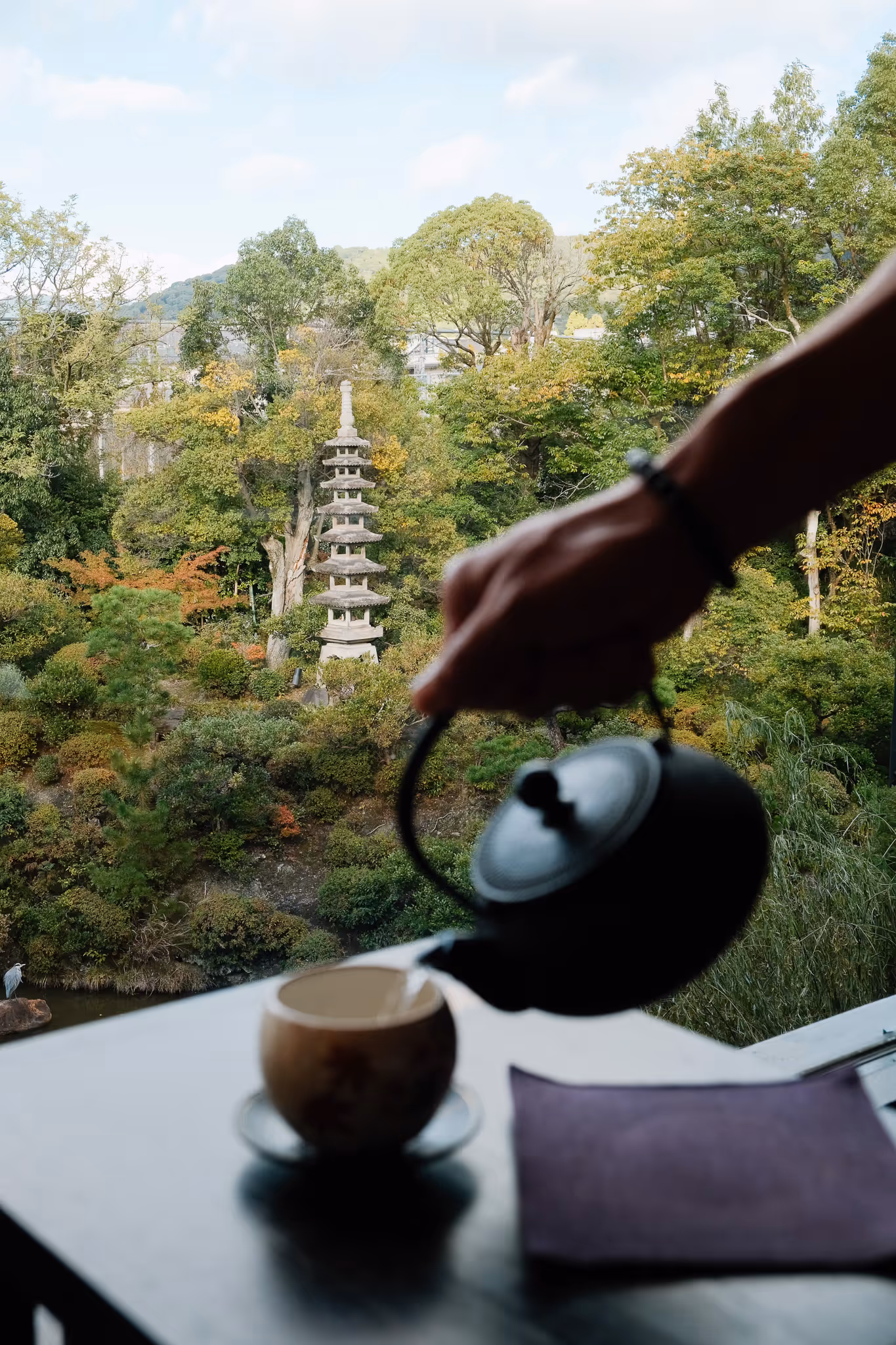Hand pouring tea from a black teapot into a cup with a Japanese garden featuring a stone pagoda in the background.