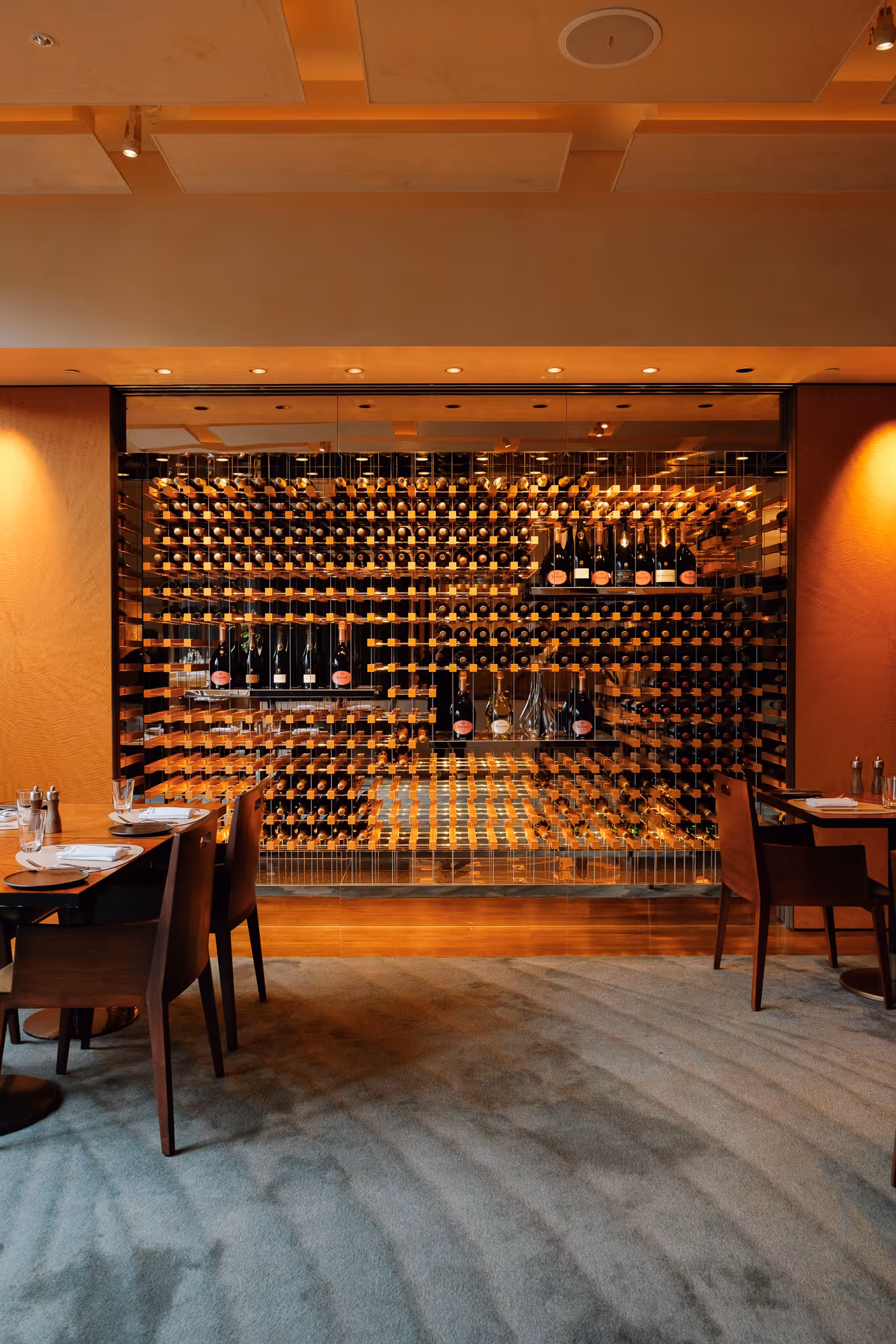 Elegant restaurant interior with a large glass-enclosed wine rack filled with bottles, wooden tables and chairs set with plates and glasses, and warm ambient lighting.