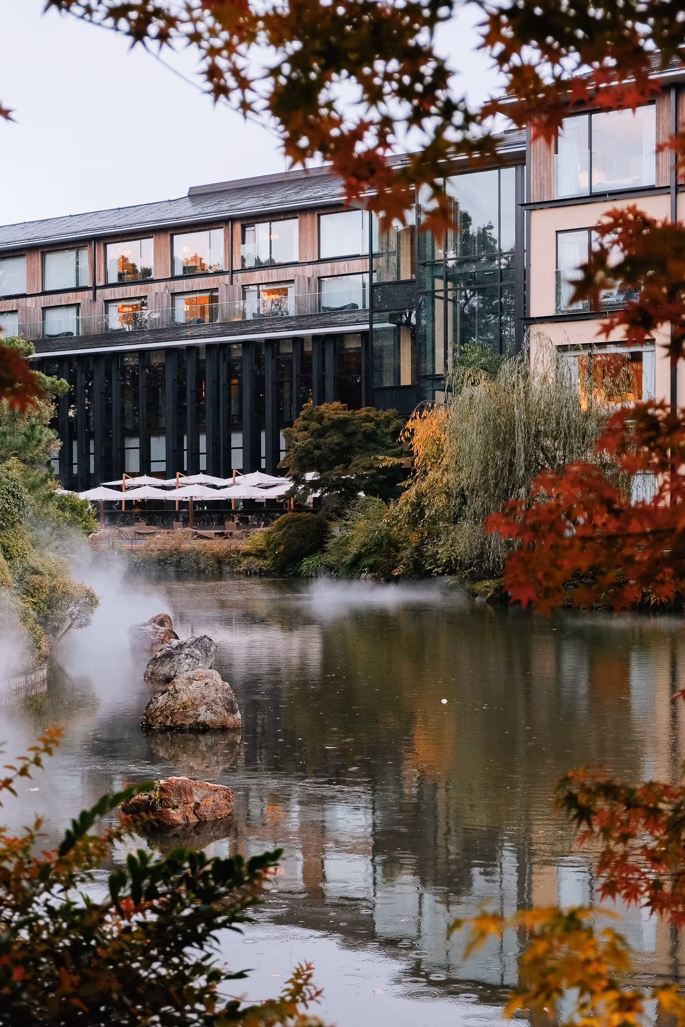 Modern building with large windows behind a calm pond surrounded by autumn foliage and misty rocks.
