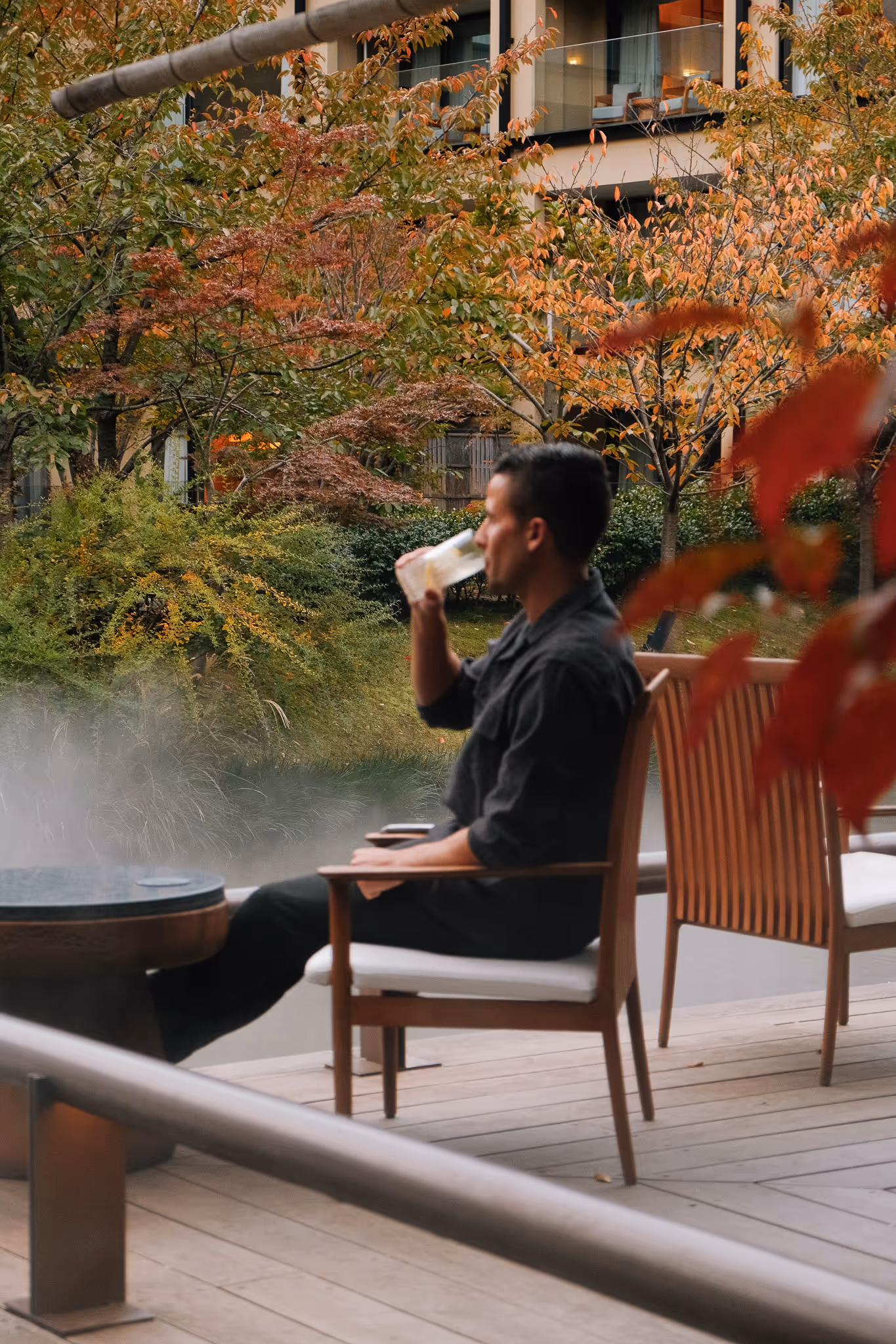 Interior of a wooden Japanese-style room with wooden chairs, tables, and a large circular ceiling light, overlooking a garden through glass sliding doors.
