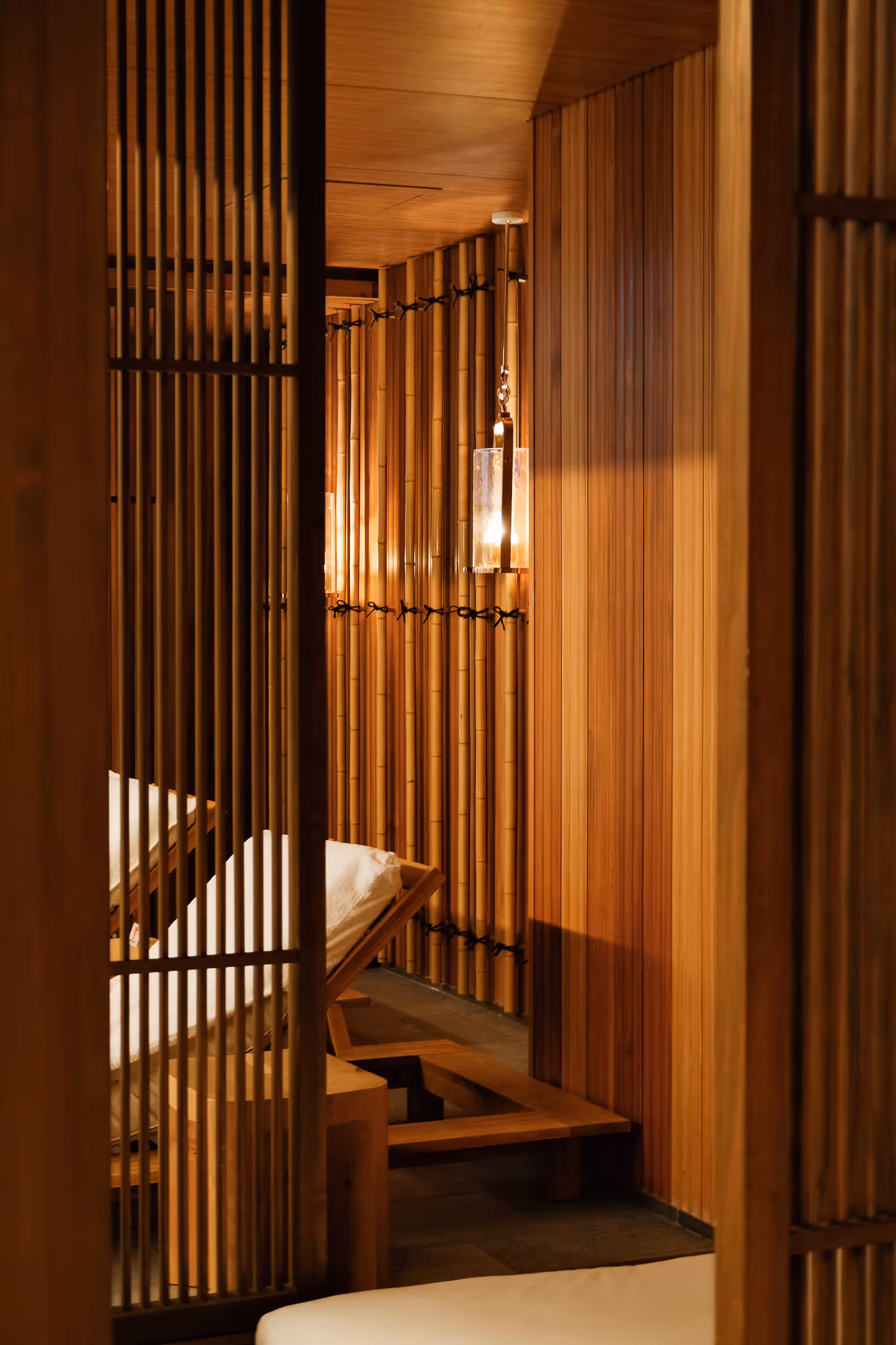 Warmly lit wooden interior with vertical slats, bamboo wall, cushioned lounge chairs, and a hanging lantern.