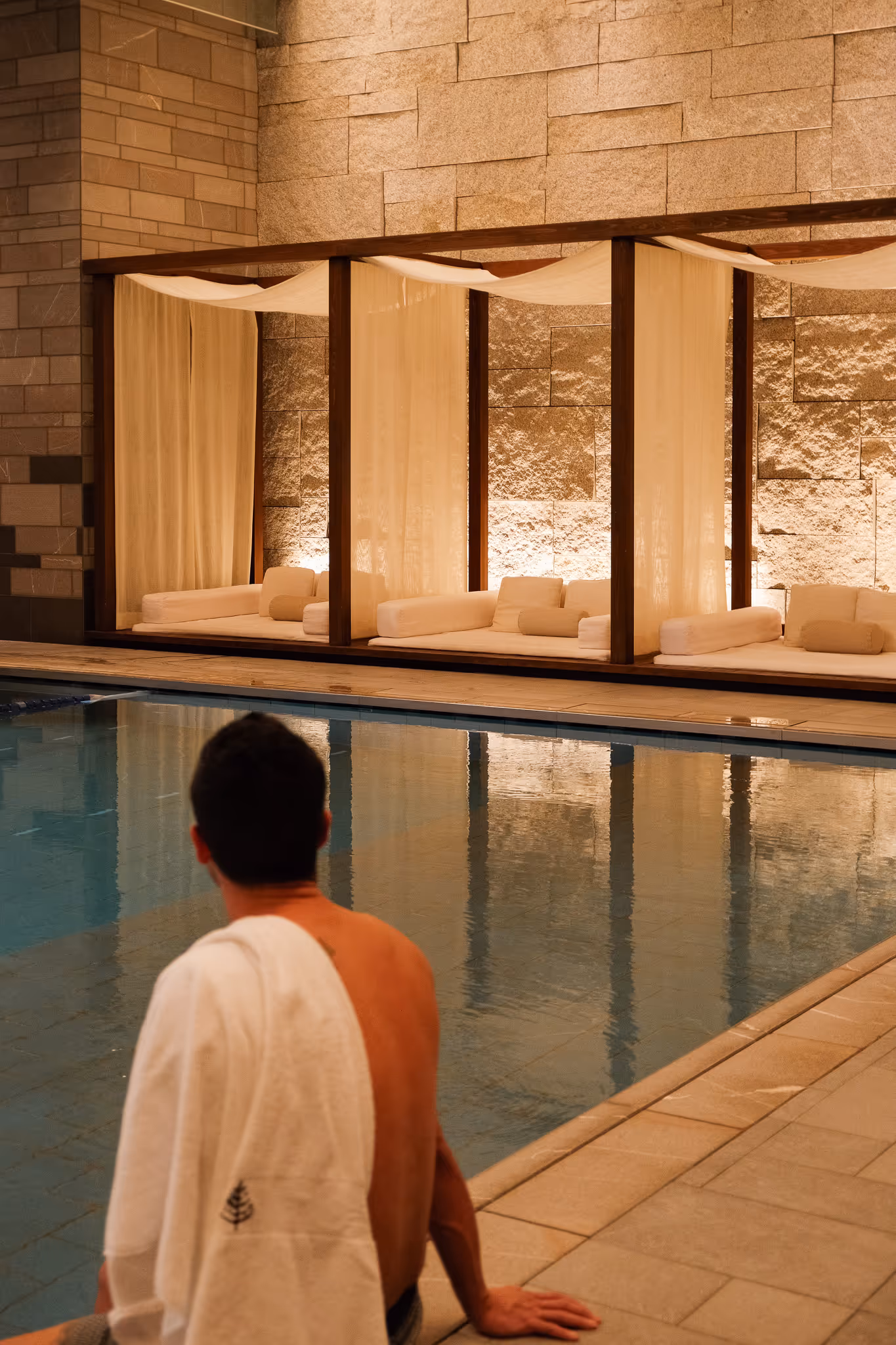 Man with a towel over his shoulder sitting at the edge of an indoor pool facing three lounge beds with sheer curtains and warm lighting.
