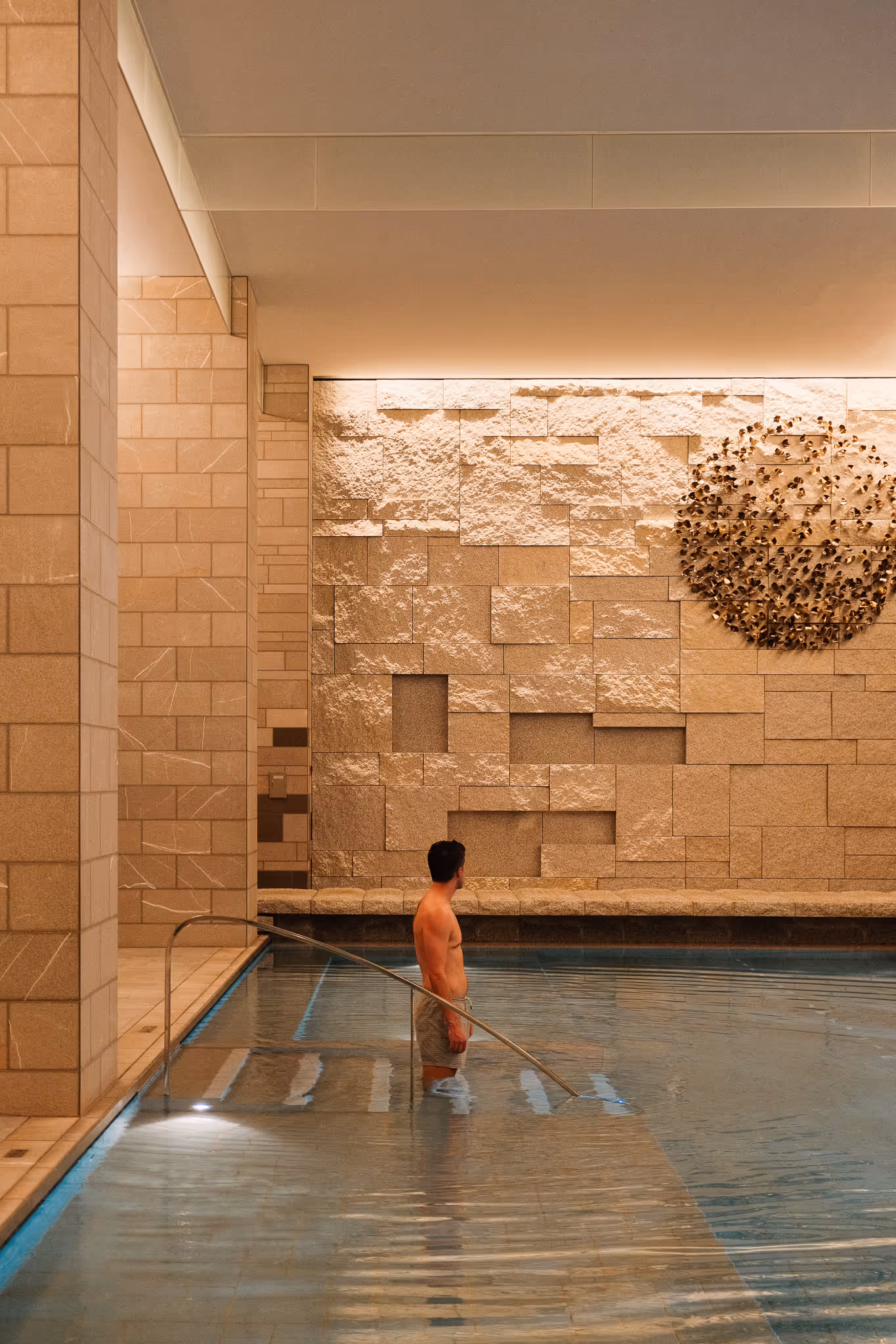 Man standing waist-deep in an indoor pool next to stone walls with decorative metal artwork.