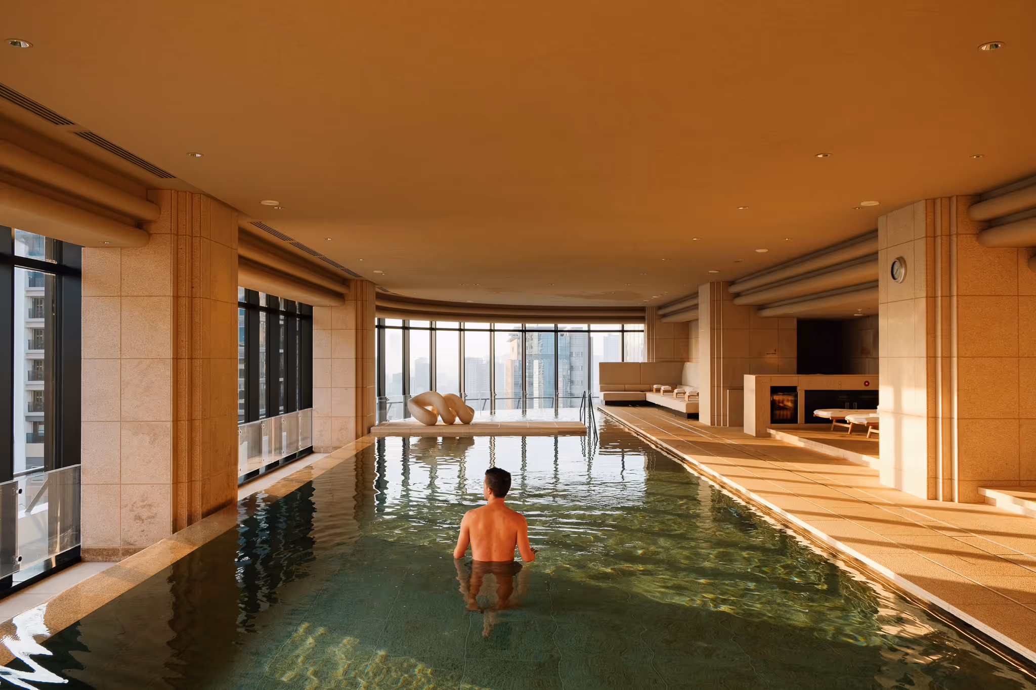 Man standing in an indoor pool with floor-to-ceiling windows overlooking a city skyline.