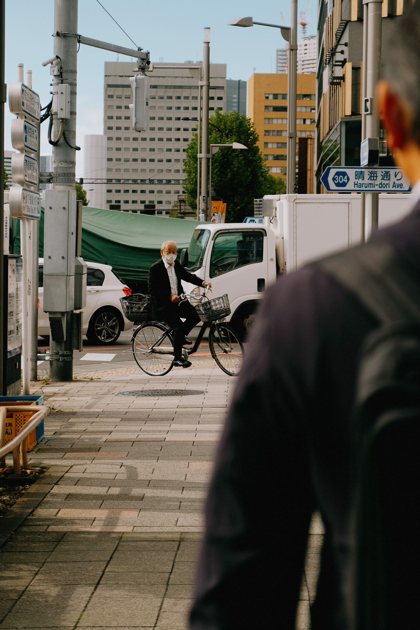 Elderly man wearing a mask riding a bicycle on an urban sidewalk with buildings and vehicles in the background.